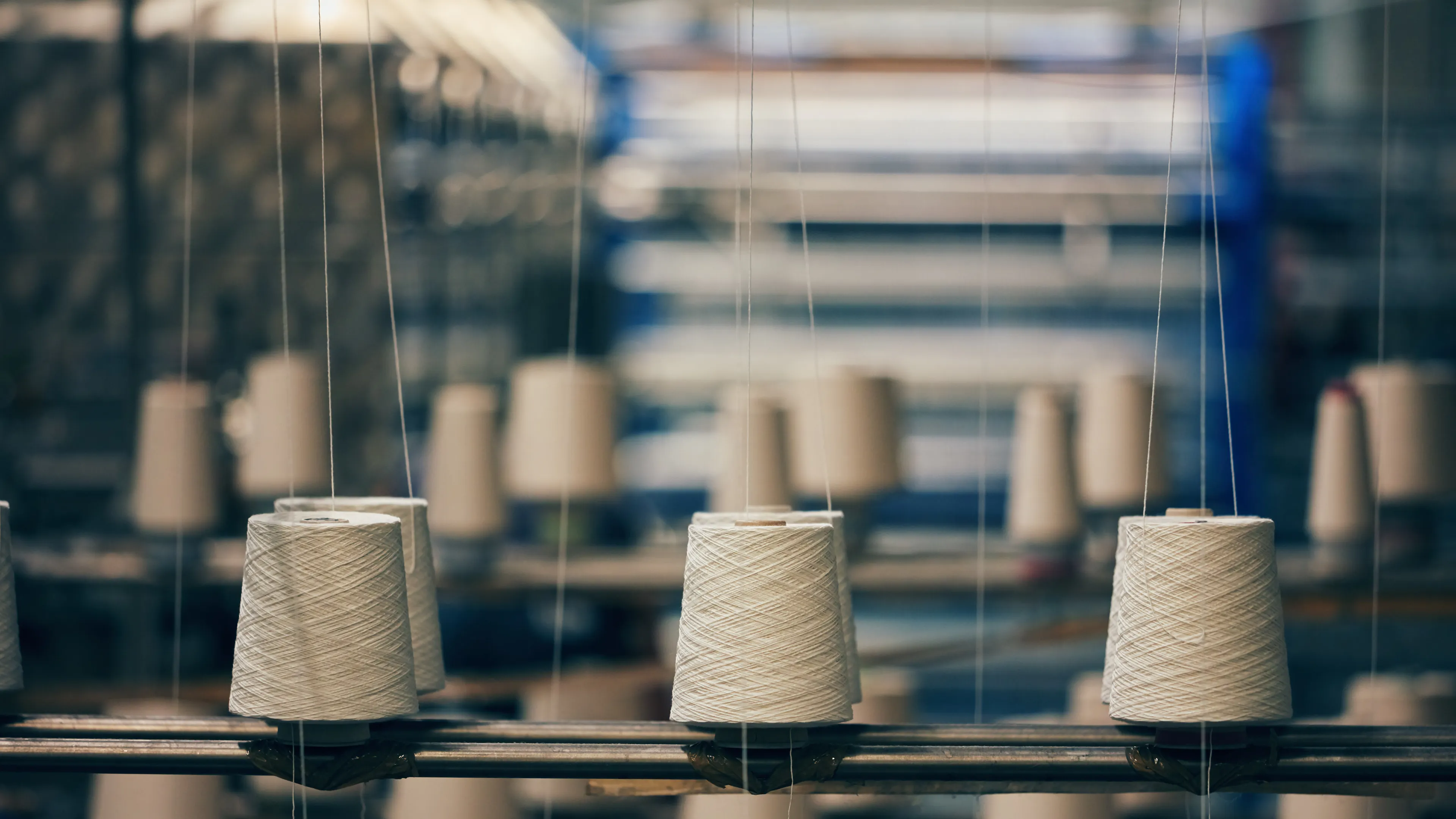 Spools of white thread are neatly arranged on a textile manufacturing machine in a factory setting. The focus is on the spools, with blurred machinery and industrial equipment in the background. The scene suggests a production process in progress.