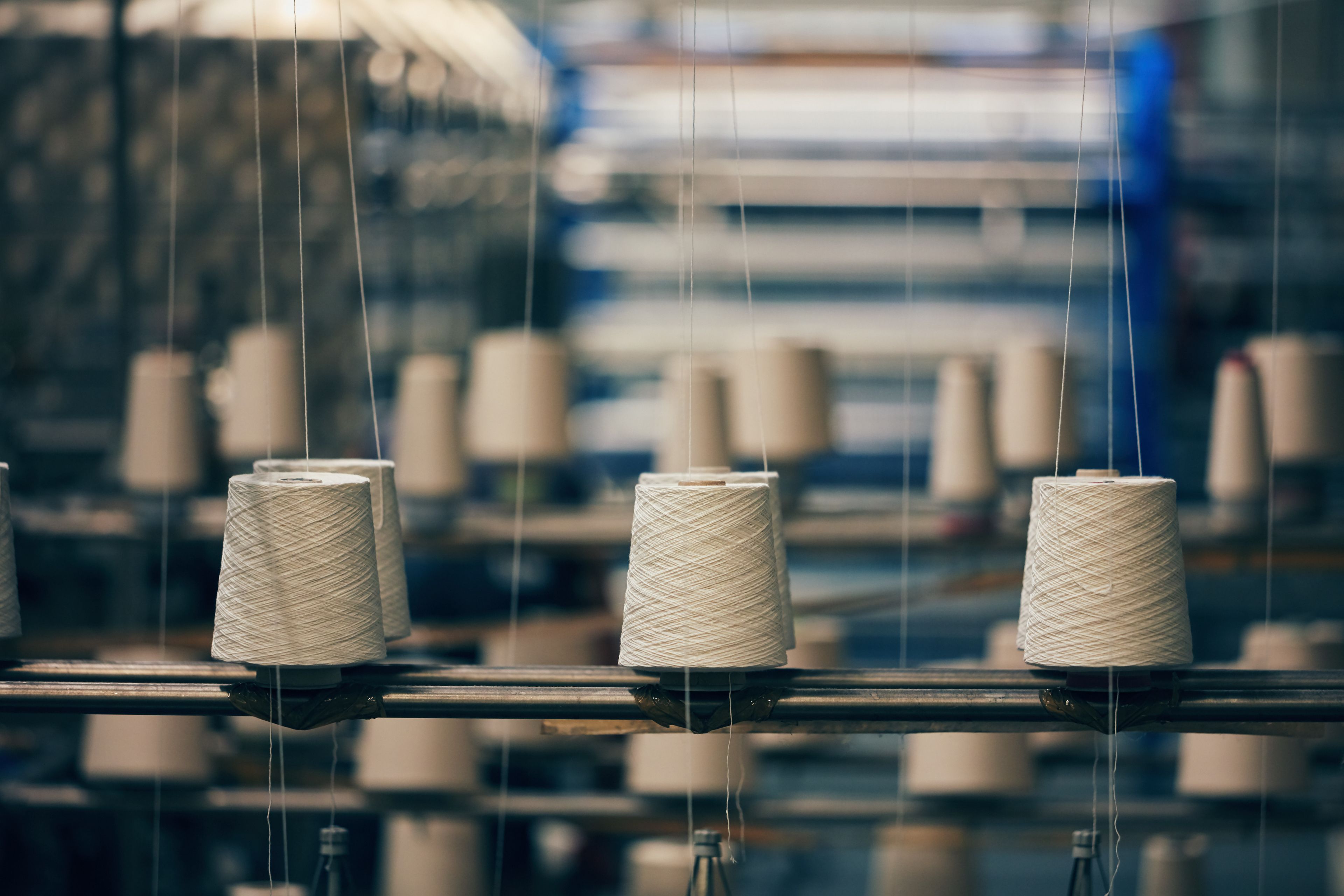 Spools of white thread are neatly arranged on a textile manufacturing machine in a factory setting. The focus is on the spools, with blurred machinery and industrial equipment in the background. The scene suggests a production process in progress.