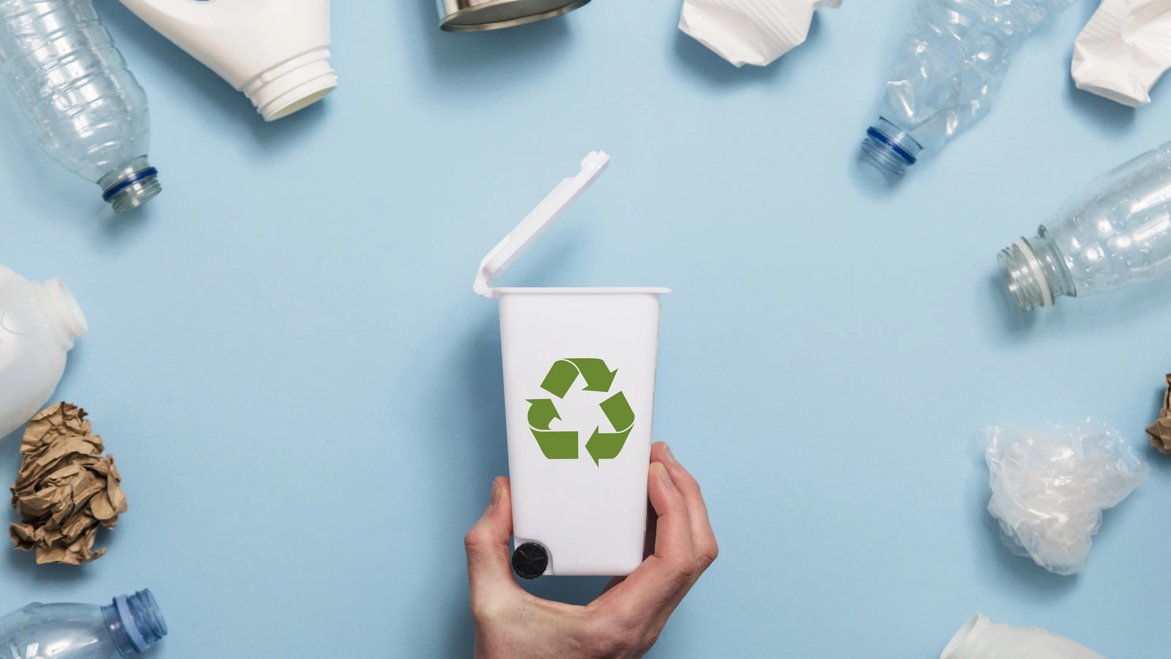 A hand holds a small white recycling bin with a green recycling symbol, surrounded by various recyclable materials like plastic bottles, cans, and paper on a light blue background. The bin’s lid is open, suggesting disposal of items. The image emphasizes recycling and environmental awareness.