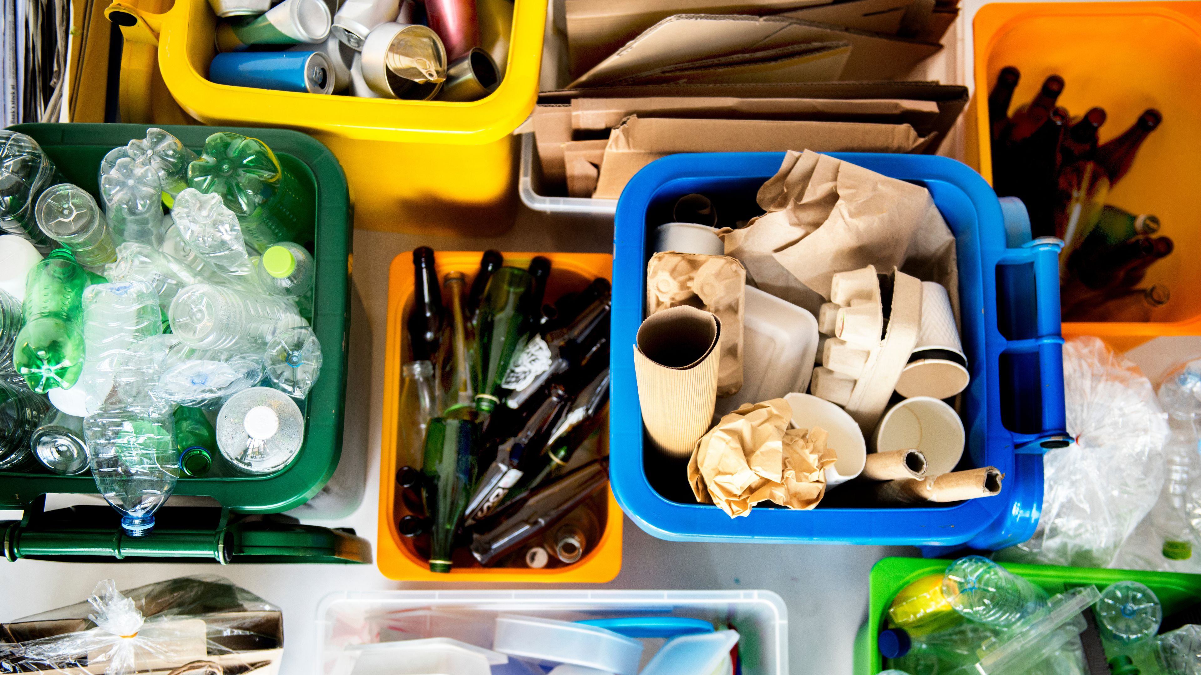 Various containers filled with recyclable materials, such as plastic bottles, glass bottles, cardboard, paper, and aluminum cans. Each bin is color-coded, suggesting organized recycling or waste sorting. The image promotes sustainability and responsible waste management.