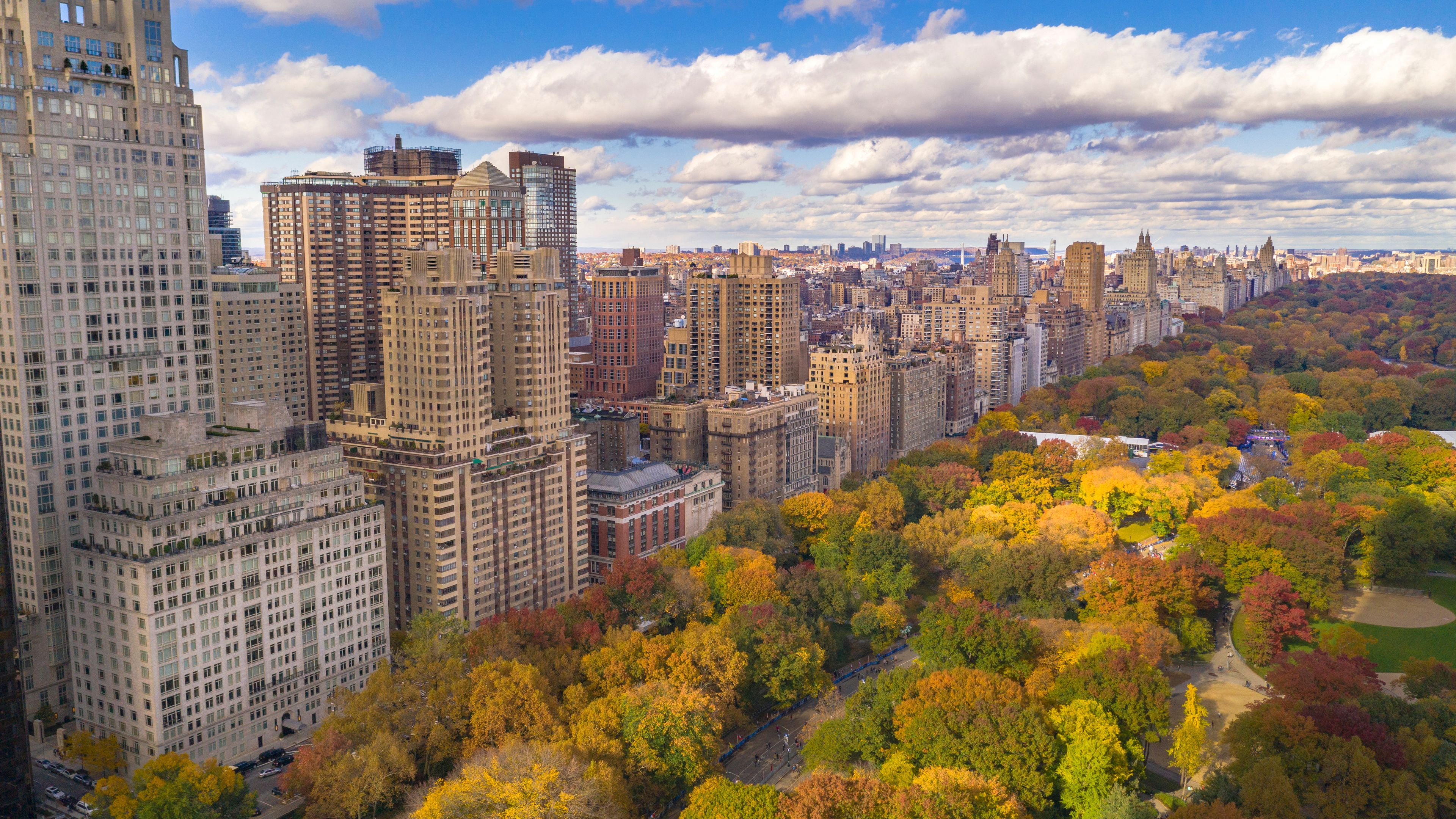 Aerial view of Manhattan's Upper East Side with Central Park in the foreground during autumn. The colorful foliage contrasts with the tall residential buildings and a partly cloudy sky above. The park's pathways and open spaces are visible amidst the vibrant trees.