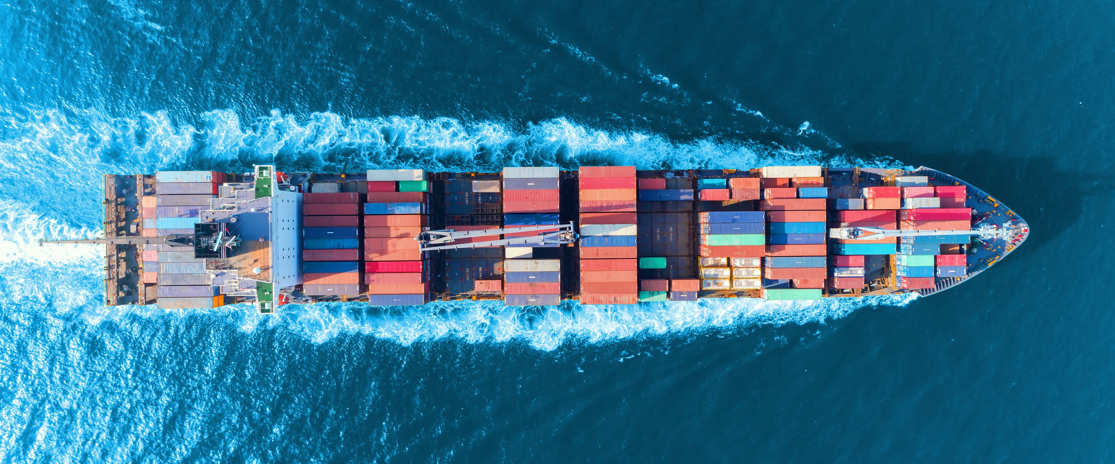 Aerial view of a large cargo ship loaded with multicolored shipping containers sailing through deep blue ocean water. The ship creates white waves as it moves forward. The scene conveys transportation and global trade.