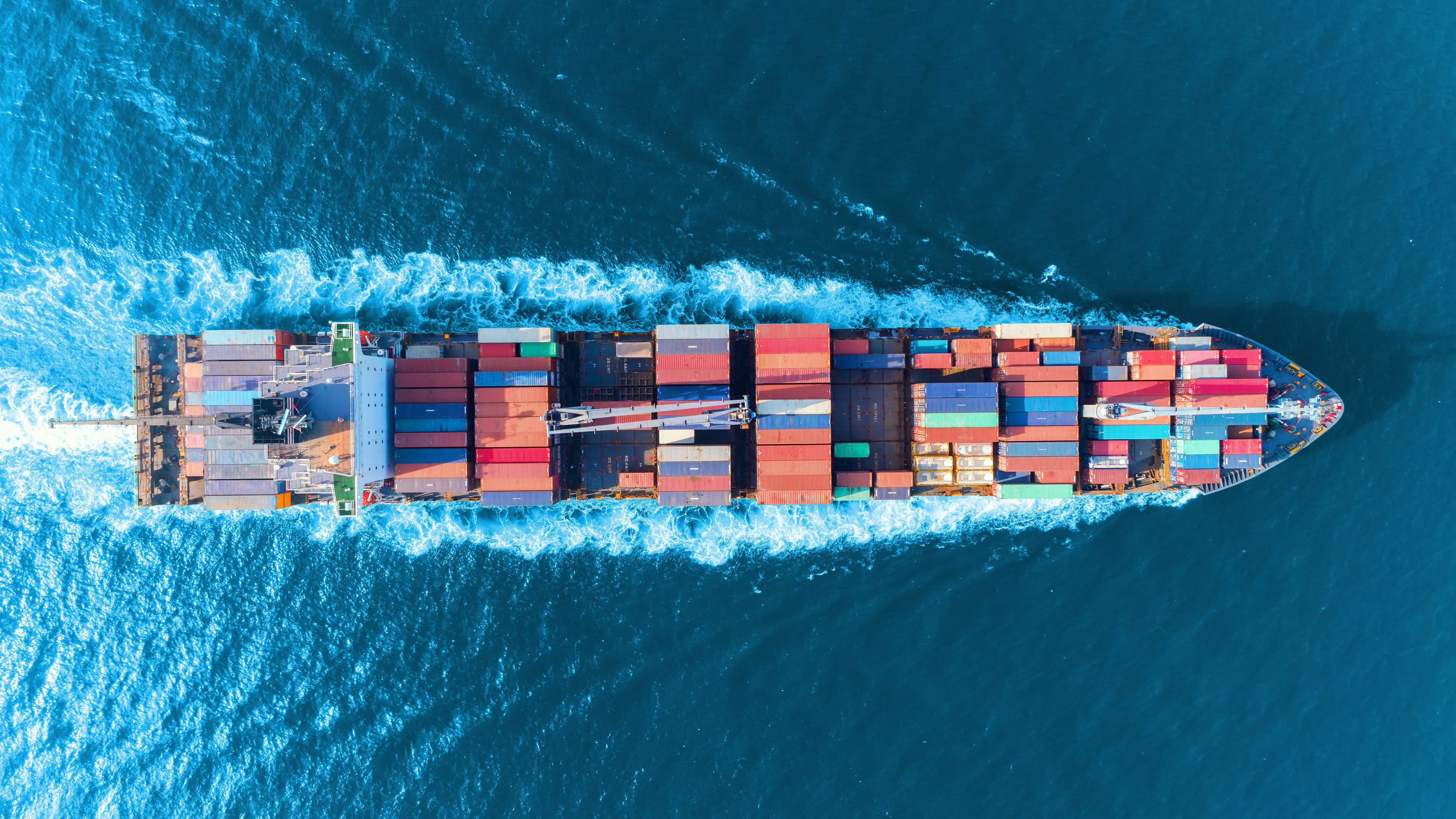 Aerial view of a large cargo ship loaded with multicolored shipping containers sailing through deep blue ocean water. The ship creates white waves as it moves forward. The scene conveys transportation and global trade.
