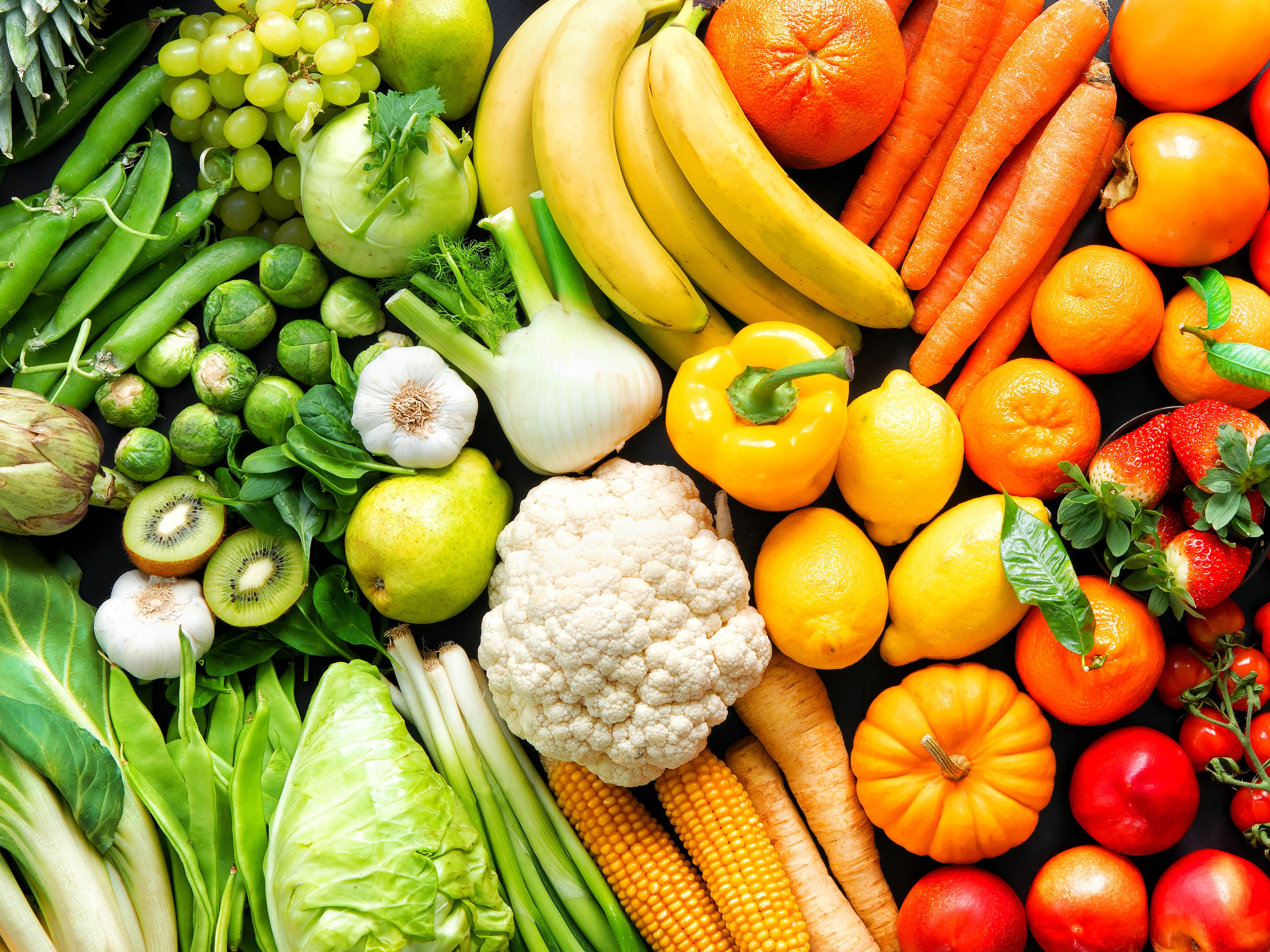 An array of fresh fruits and vegetables arranged in a rainbow gradient, starting with green leafy produce on the left and transitioning through yellow, orange, red, and purple items to the right. It includes lettuce, bananas, oranges, peppers, eggplants, and grapes. The vibrant colors emphasize the diversity of fresh, healthy produce.
