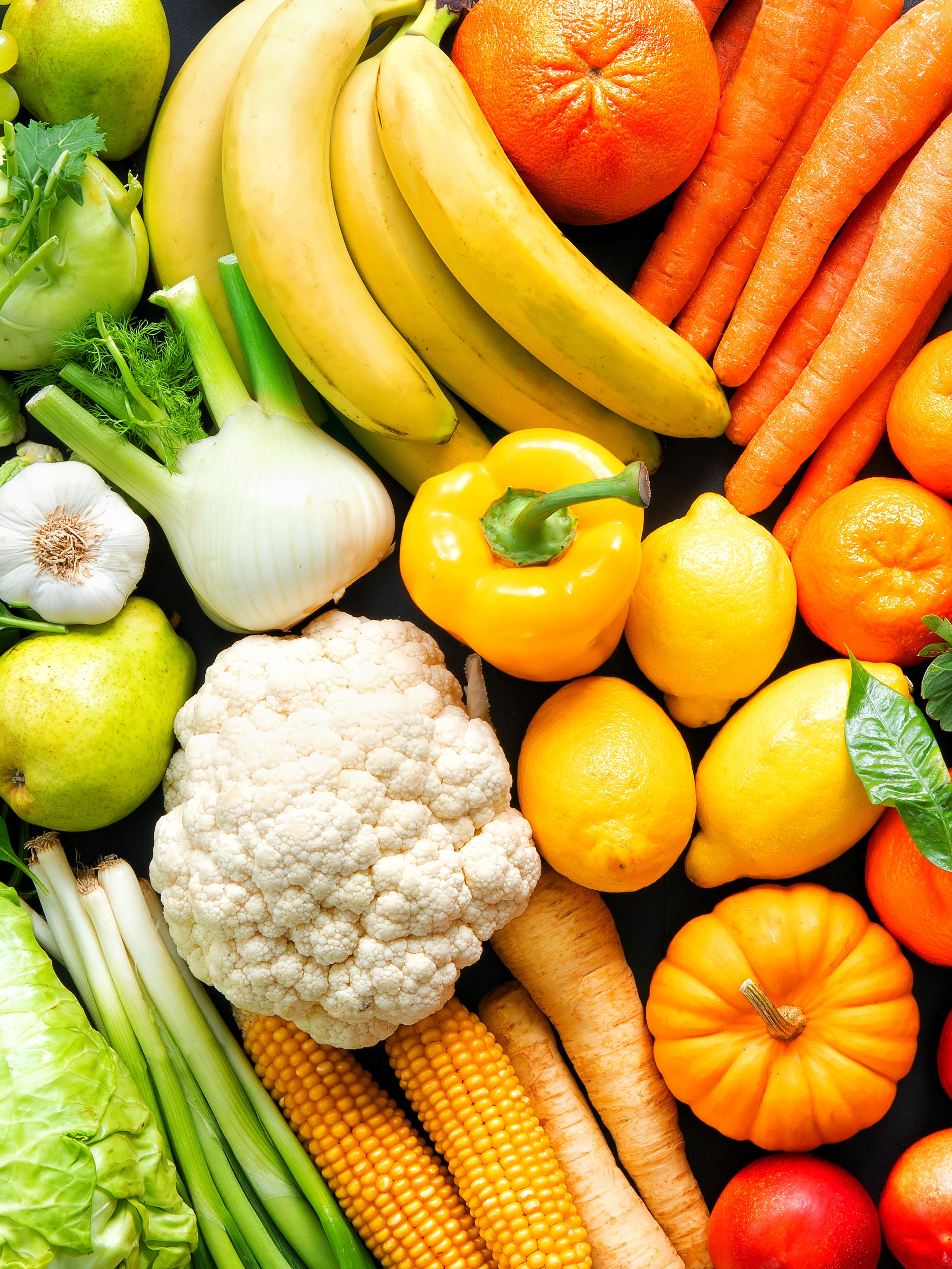 An array of fresh fruits and vegetables arranged in a rainbow gradient, starting with green leafy produce on the left and transitioning through yellow, orange, red, and purple items to the right. It includes lettuce, bananas, oranges, peppers, eggplants, and grapes. The vibrant colors emphasize the diversity of fresh, healthy produce.
