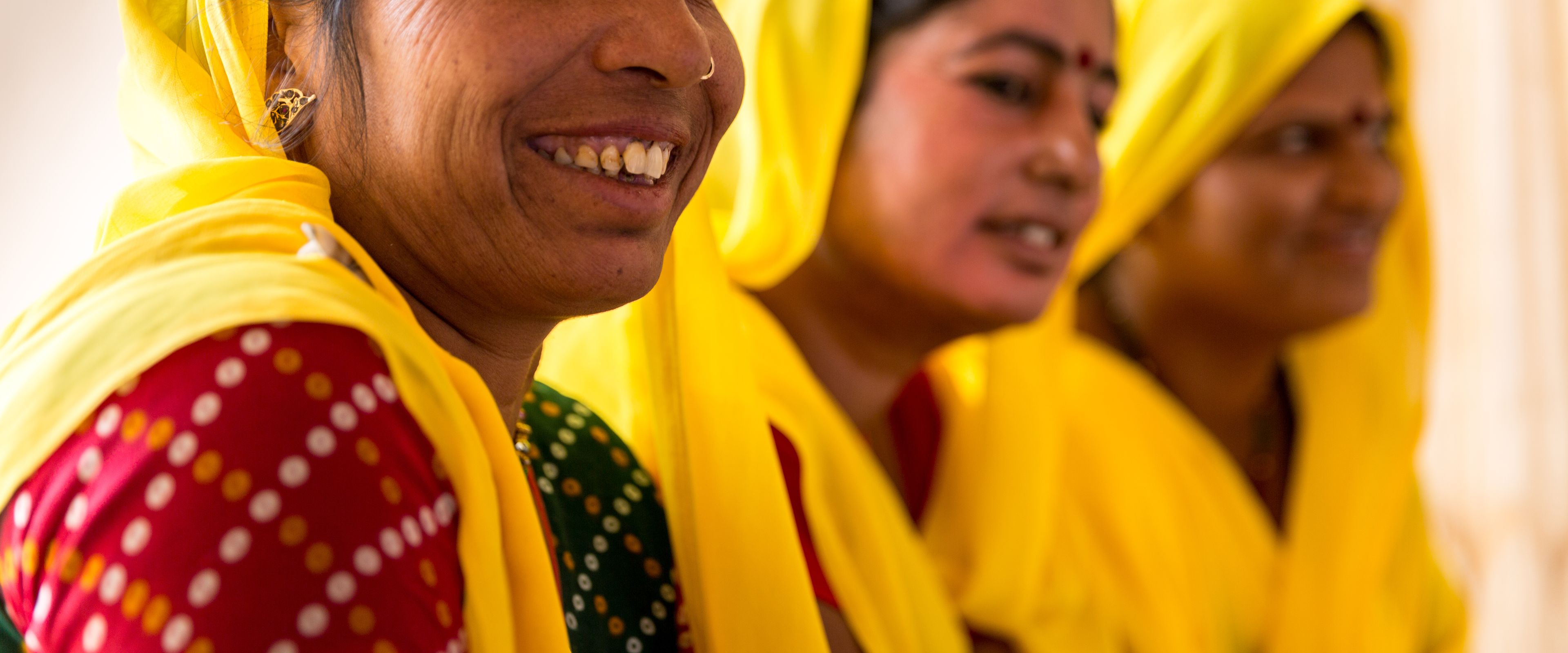 Three women dressed in vibrant yellow sarees and traditional attire are gathered together, smiling and conversing. The woman in the foreground has a joyful expression and a red bindi on her forehead. The background is softly blurred, highlighting their interaction.