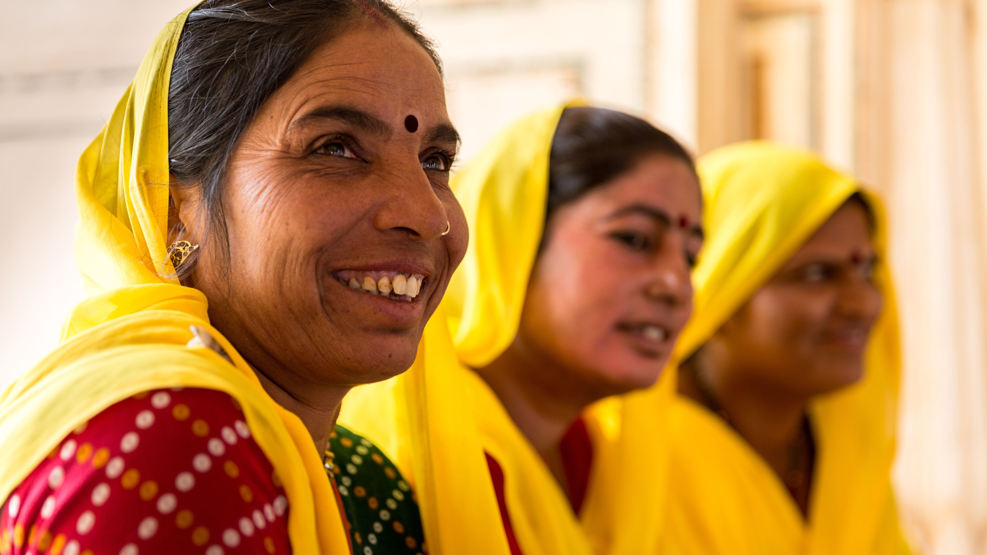 Three women dressed in vibrant yellow sarees and traditional attire are gathered together, smiling and conversing. The woman in the foreground has a joyful expression and a red bindi on her forehead. The background is softly blurred, highlighting their interaction.