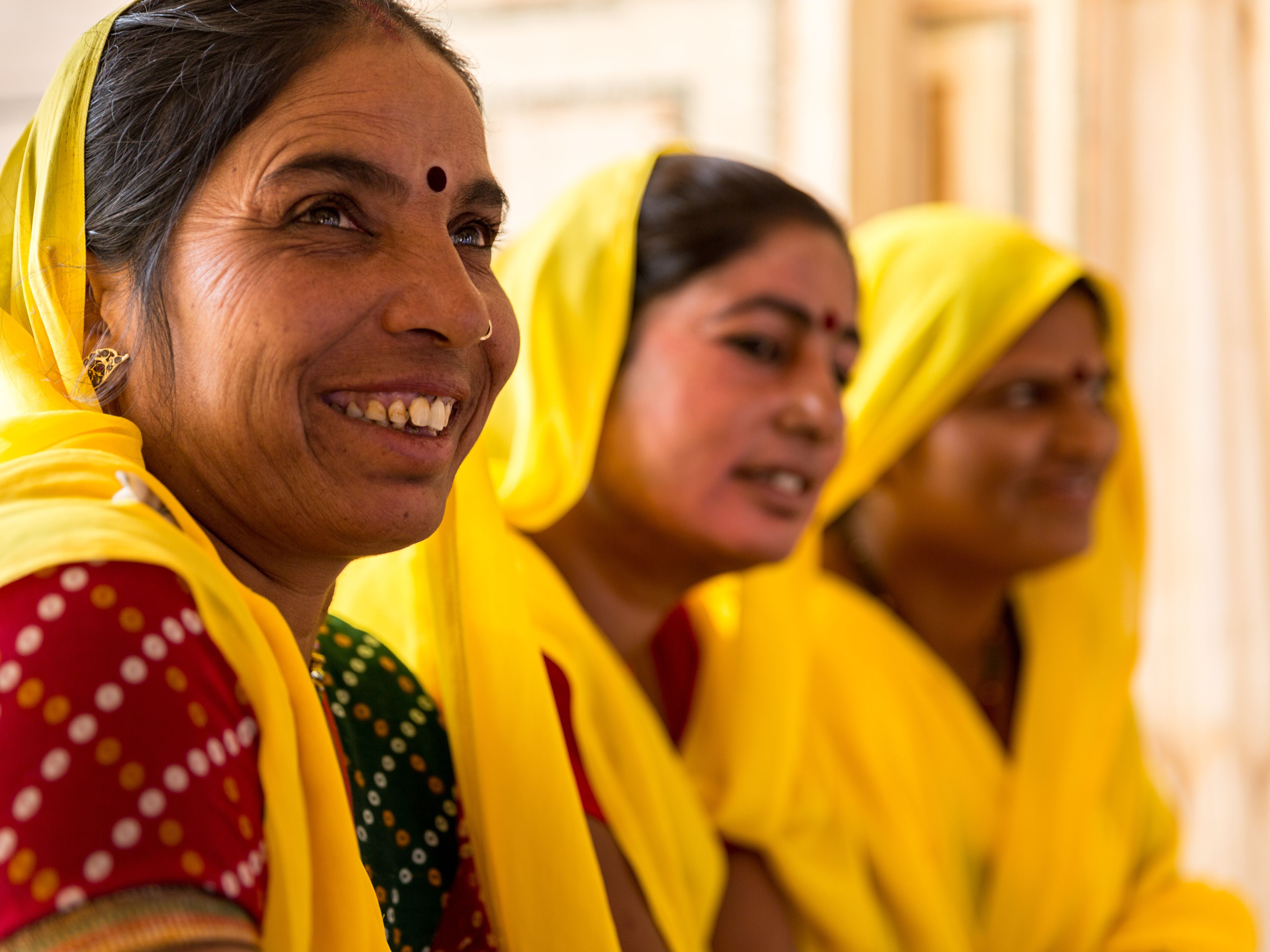 Three women dressed in vibrant yellow sarees and traditional attire are gathered together, smiling and conversing. The woman in the foreground has a joyful expression and a red bindi on her forehead. The background is softly blurred, highlighting their interaction.
