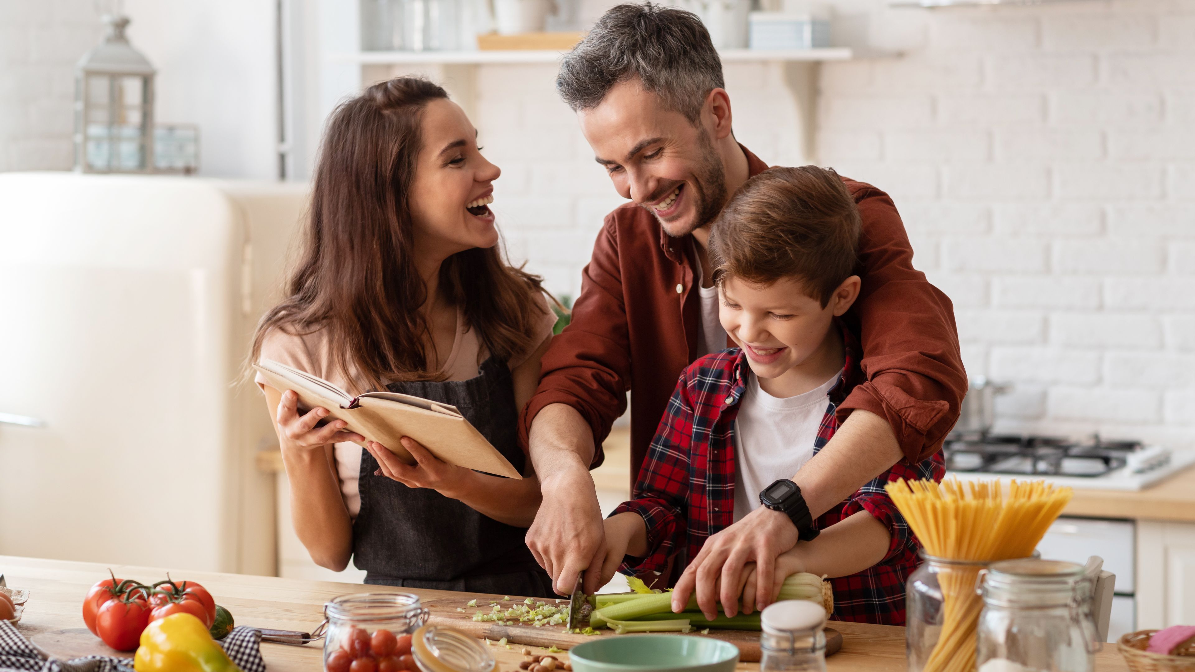 A family of three is in a kitchen preparing a meal together. The man is helping the boy chop vegetables while the woman laughs and holds a cookbook. The kitchen is bright and has various ingredients and jars on the counter.