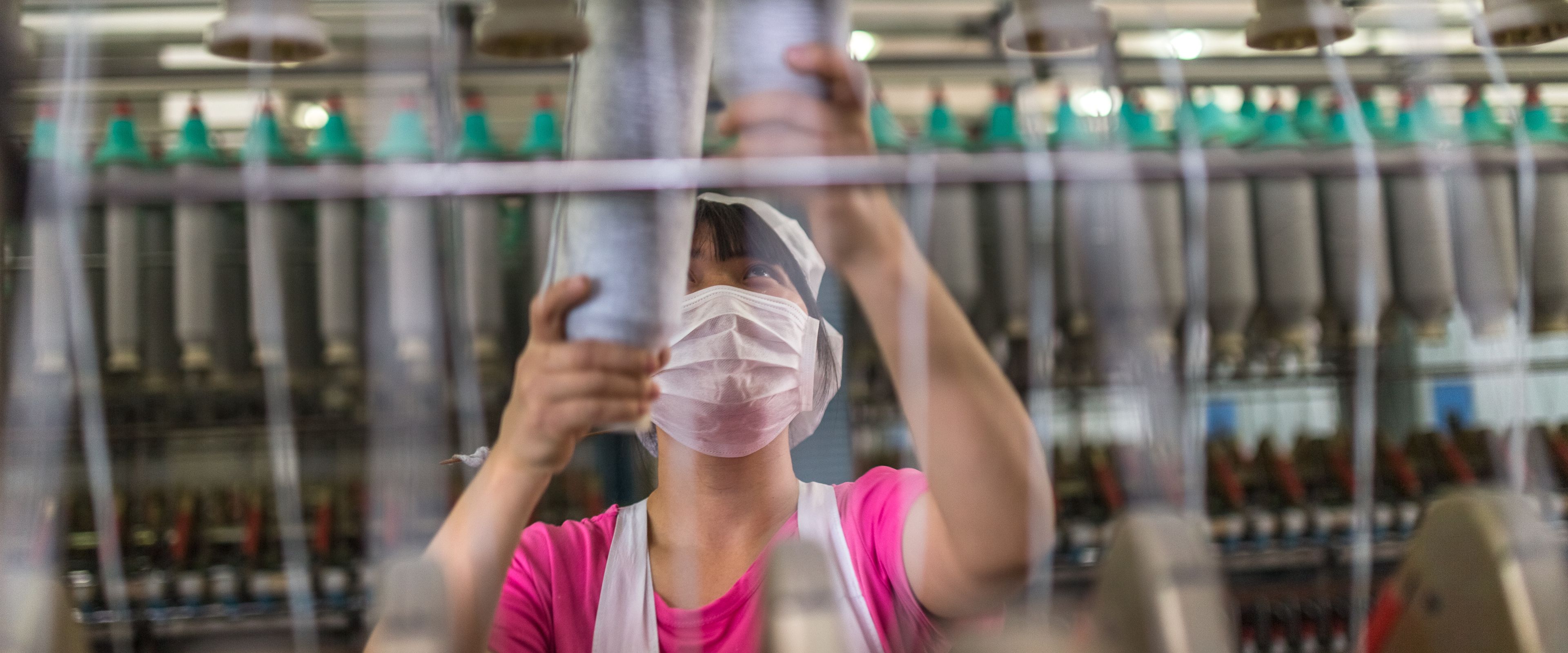 A worker wearing a face mask is arranging spools of thread on a textile machine in a factory. Rows of yarn spools and machinery are visible in the background. The scene depicts textile manufacturing processes.