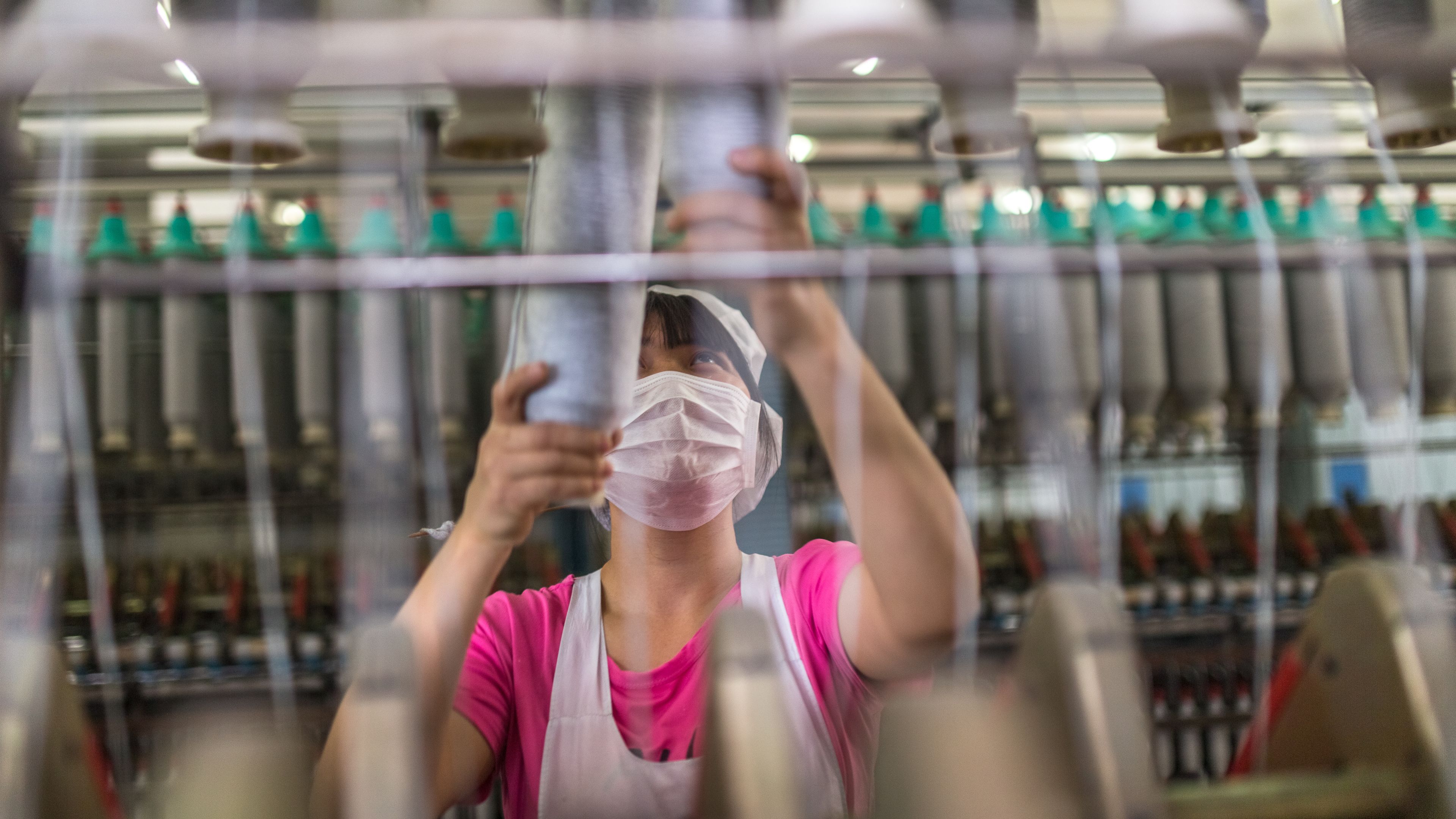 A worker wearing a face mask is arranging spools of thread on a textile machine in a factory. Rows of yarn spools and machinery are visible in the background. The scene depicts textile manufacturing processes.