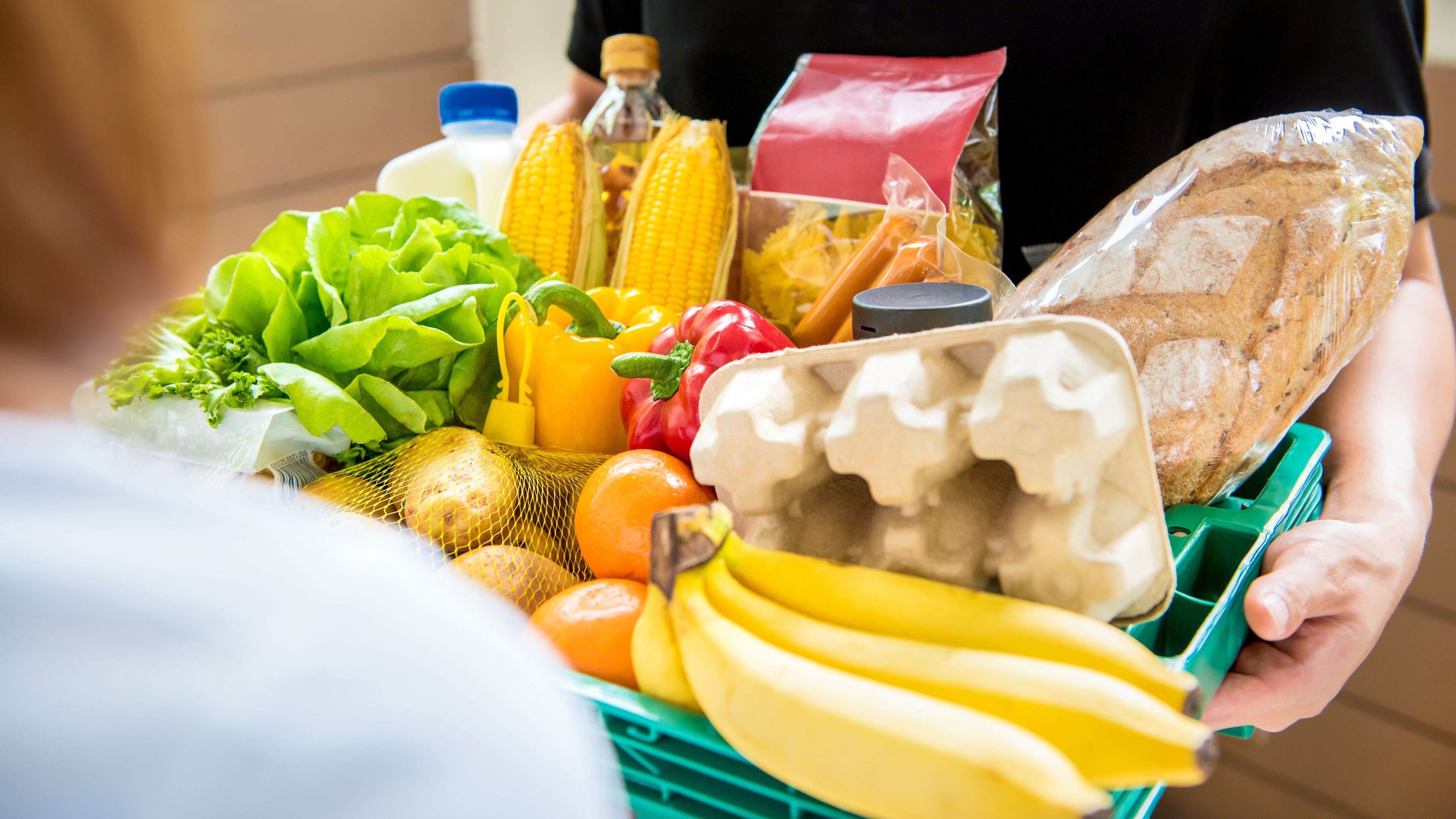 A person is holding a green crate filled with groceries, including fresh produce like bananas, oranges, lettuce, corn, and bell peppers, as well as packaged food items, bread, and eggs. Another person is partially visible in the foreground. The setting appears to be a delivery or grocery exchange outdoors.