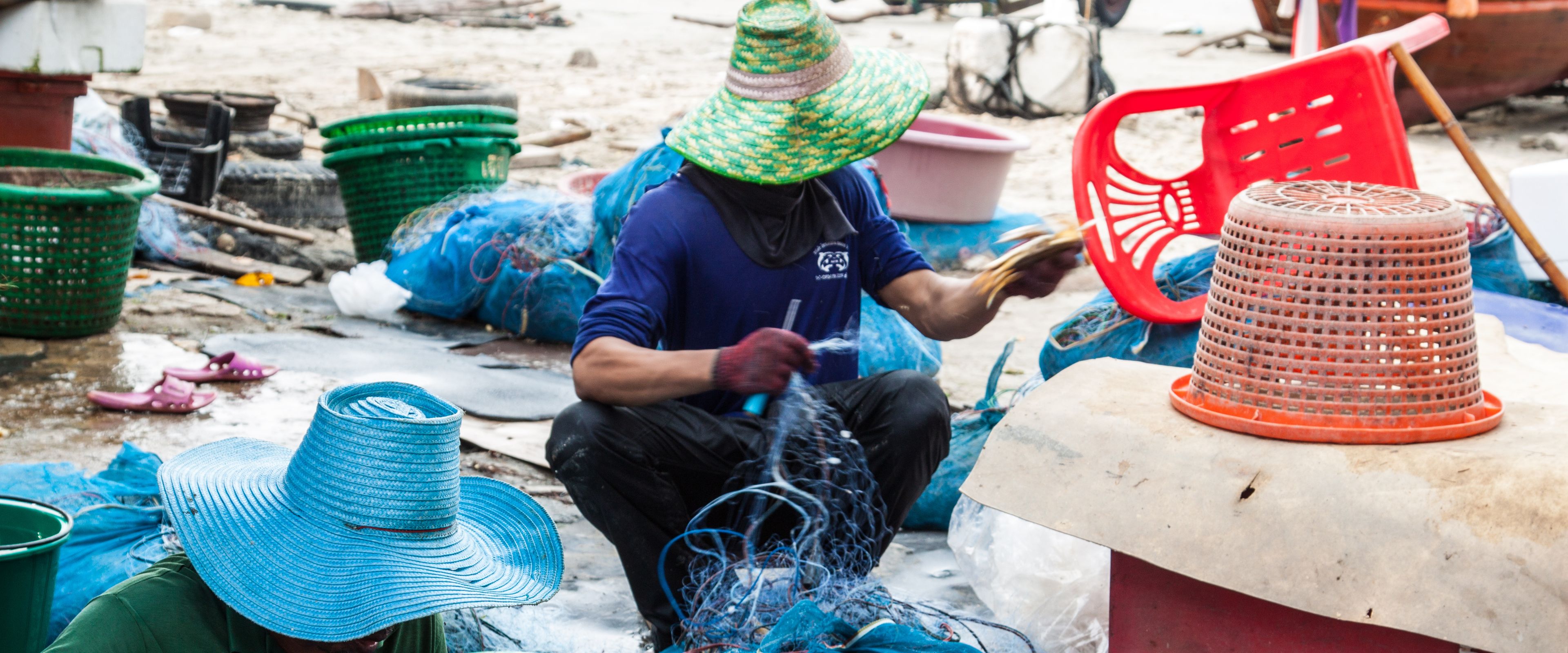 Two people wearing large, colorful straw hats sit among fishing nets and baskets, working with fishing gear. Various tools and containers surround them, indicating a fishing-related activity. The scene suggests a coastal or fishing village setting.
