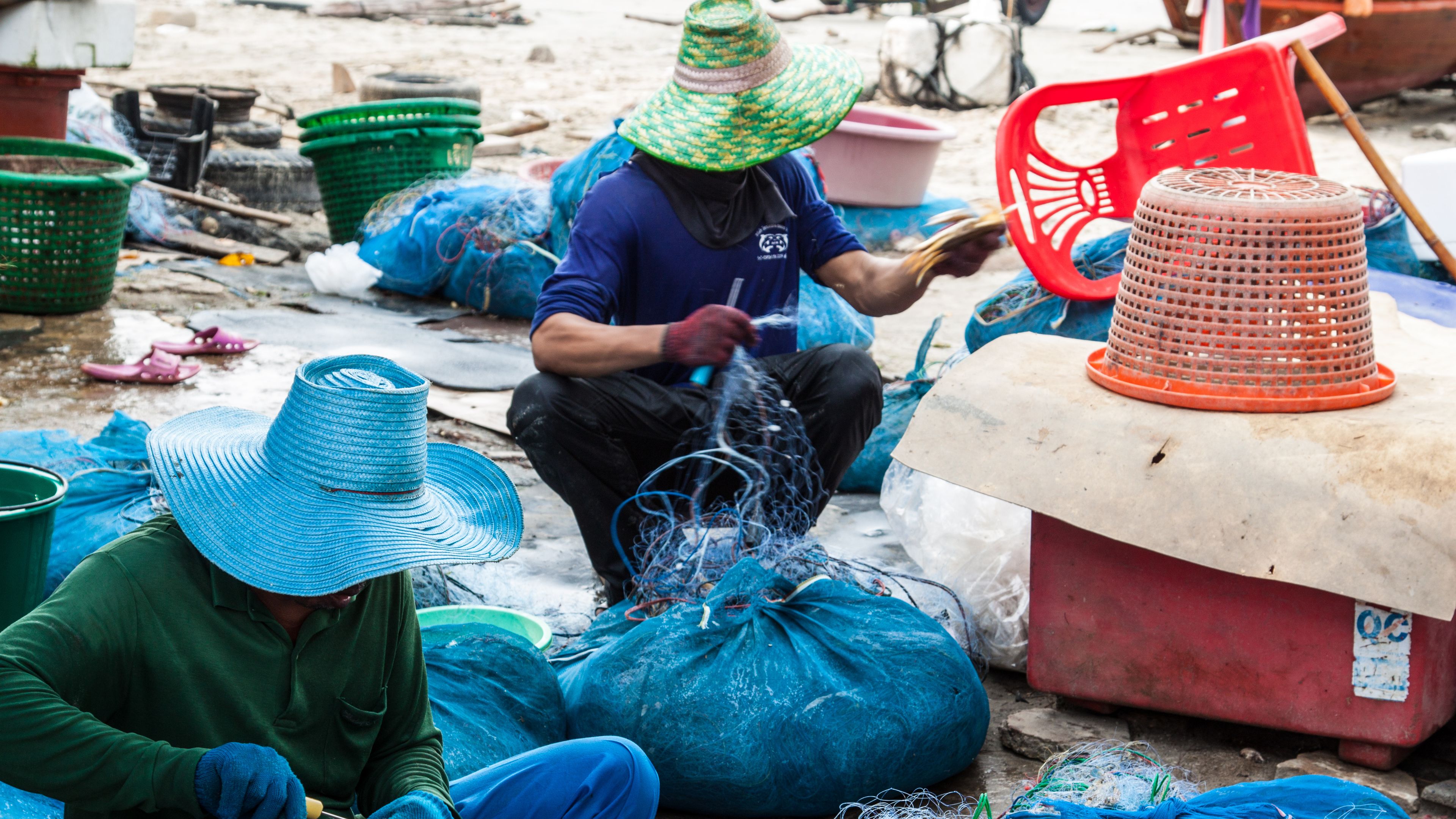 Two people wearing large, colorful straw hats sit among fishing nets and baskets, working with fishing gear. Various tools and containers surround them, indicating a fishing-related activity. The scene suggests a coastal or fishing village setting.