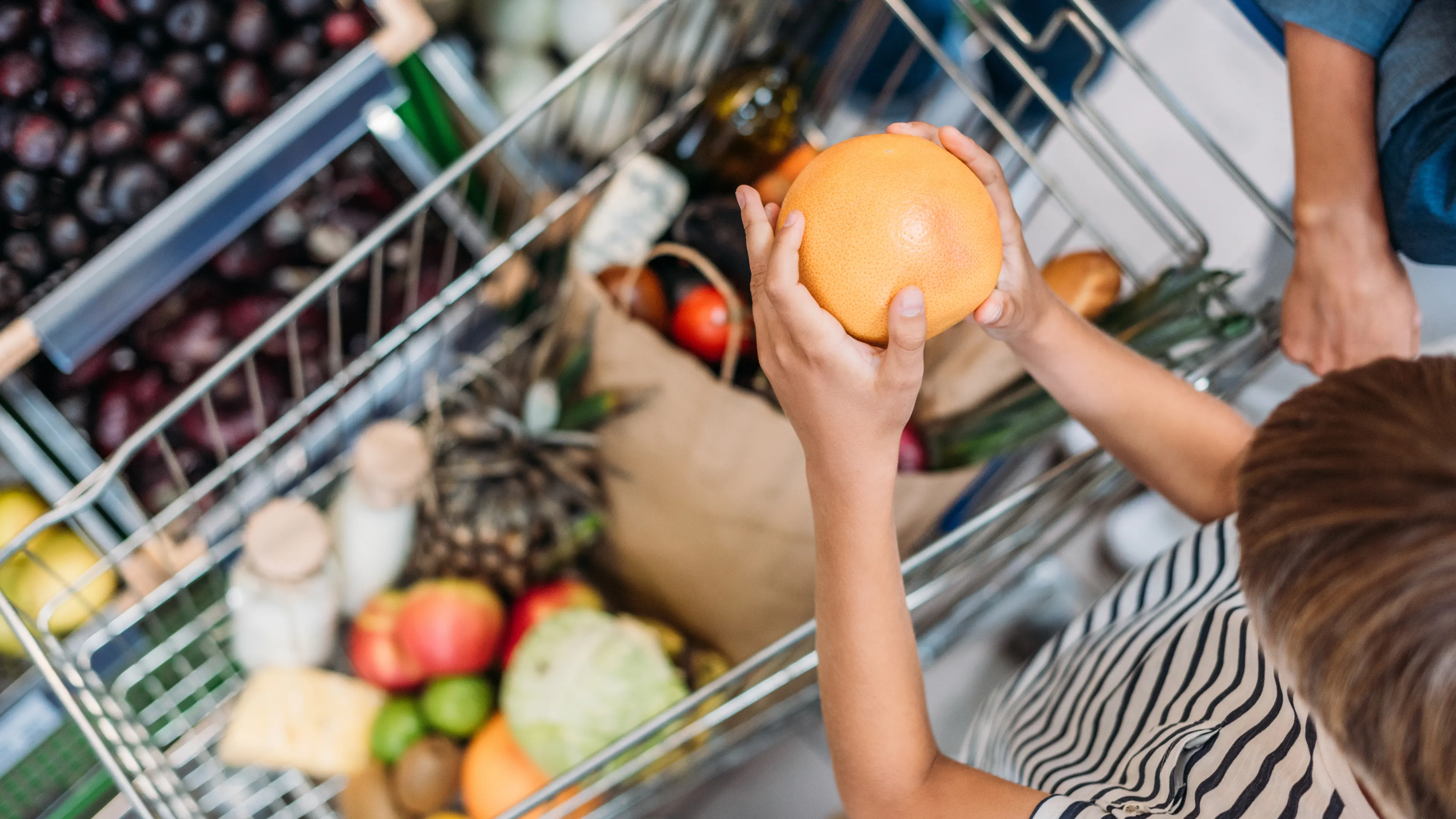 A child holding an orange stands next to a shopping cart filled with various groceries, including fruits, vegetables, and bottles. The cart is packed with colorful and fresh produce. Another person's hand is partially visible, assisting.