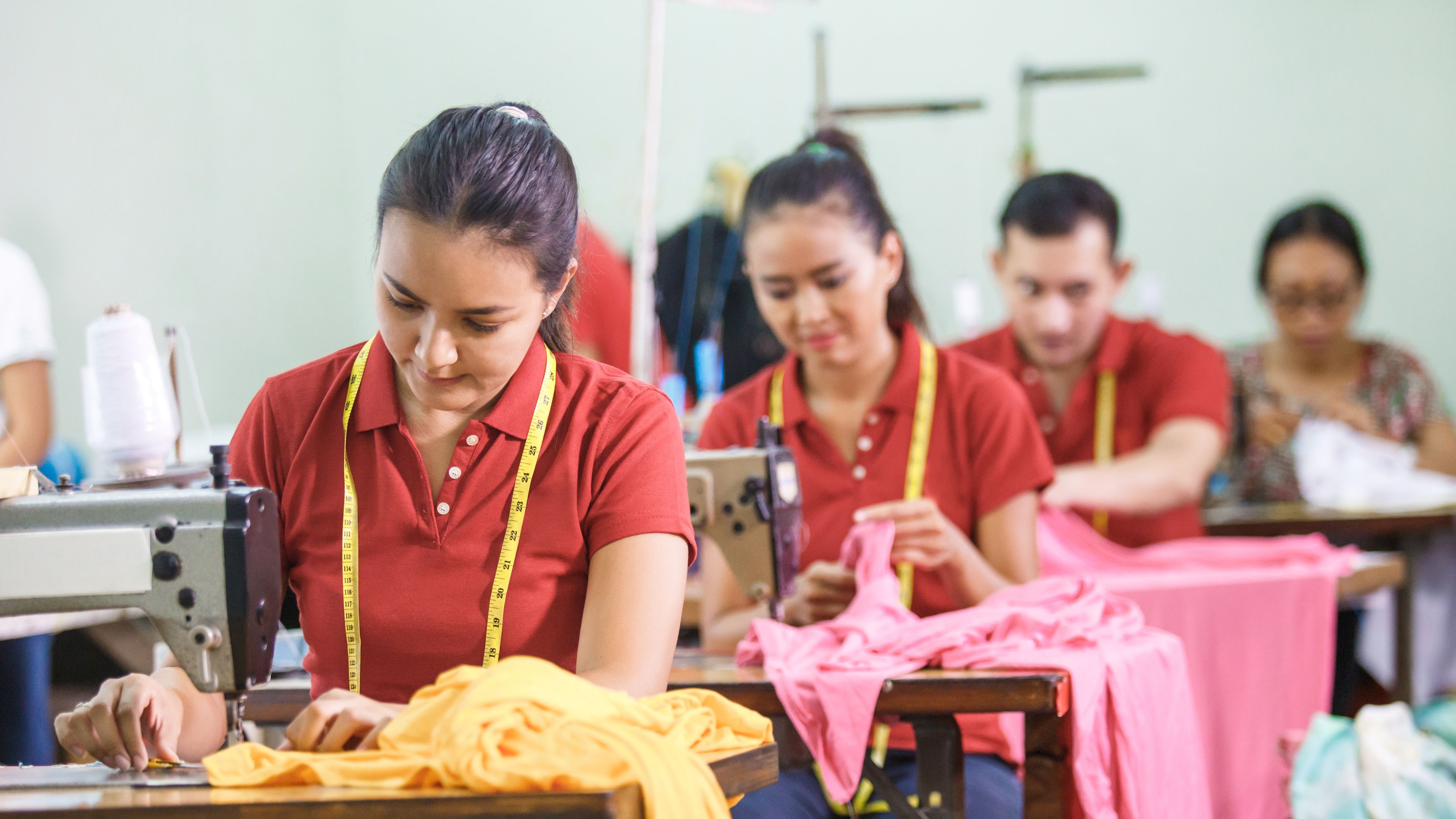 A group of people working in a garment workshop, focused on sewing and stitching colorful fabric. Two women in red shirts, wearing measuring tapes around their necks, are at sewing machines. Other workers in the background are engaged in similar tasks.