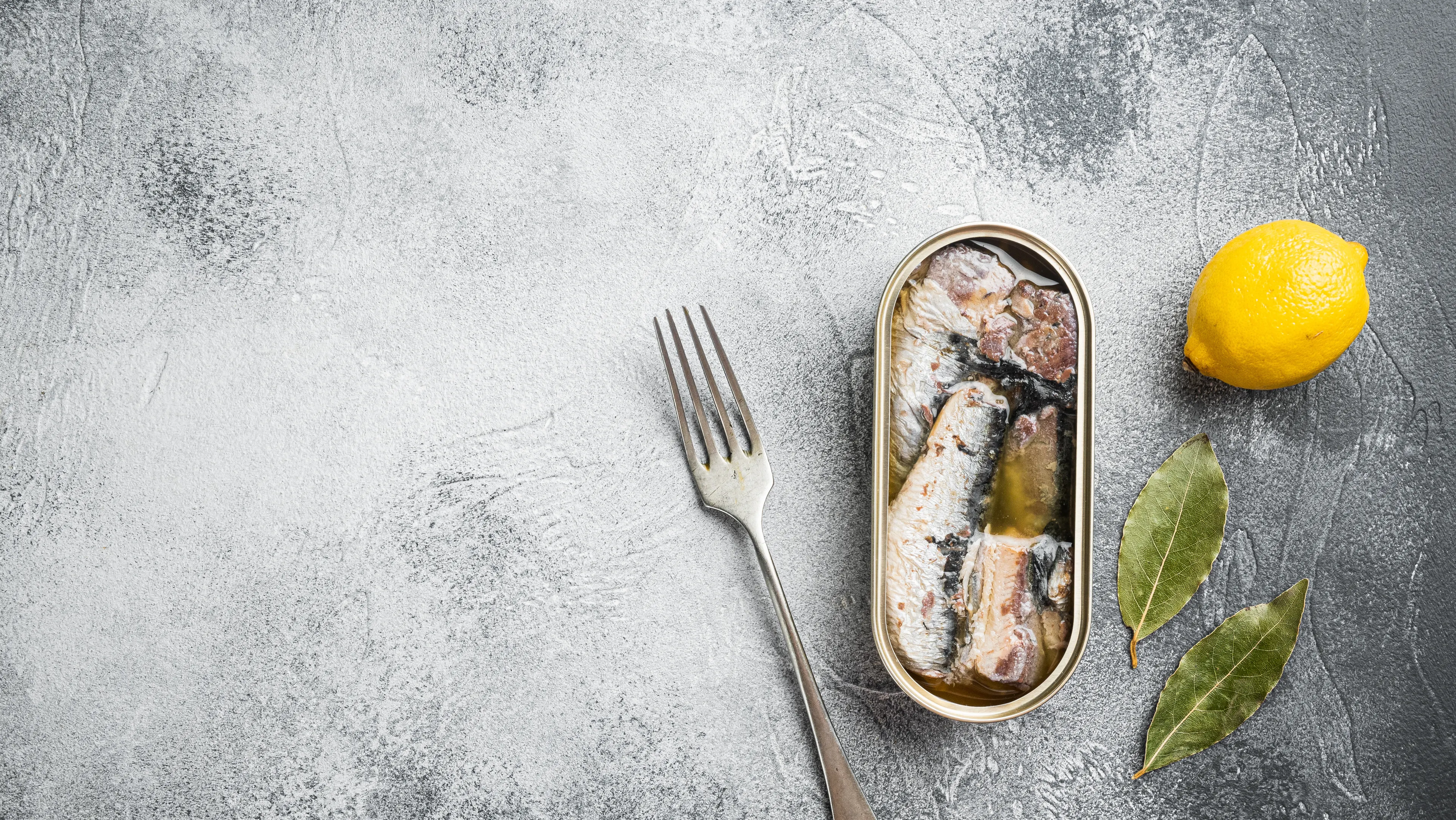 An open tin of sardines in oil is placed on a textured gray surface alongside a fork. A whole lemon and two bay leaves are arranged as garnish. The setting has a minimalist and rustic aesthetic.