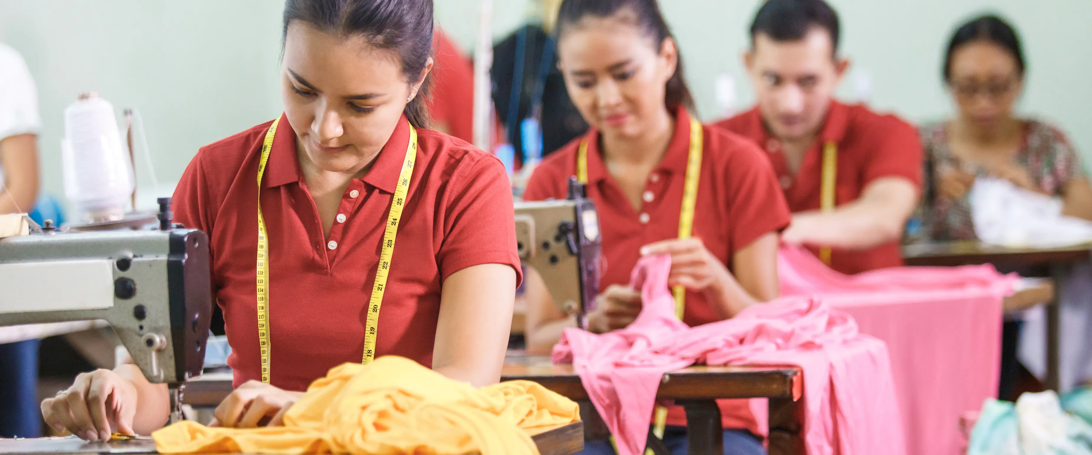 Workers in a sewing workshop are focused on stitching and handling colorful fabric. They are wearing red shirts and yellow measuring tapes around their necks. Sewing machines and fabric are visible on the shared workspace.