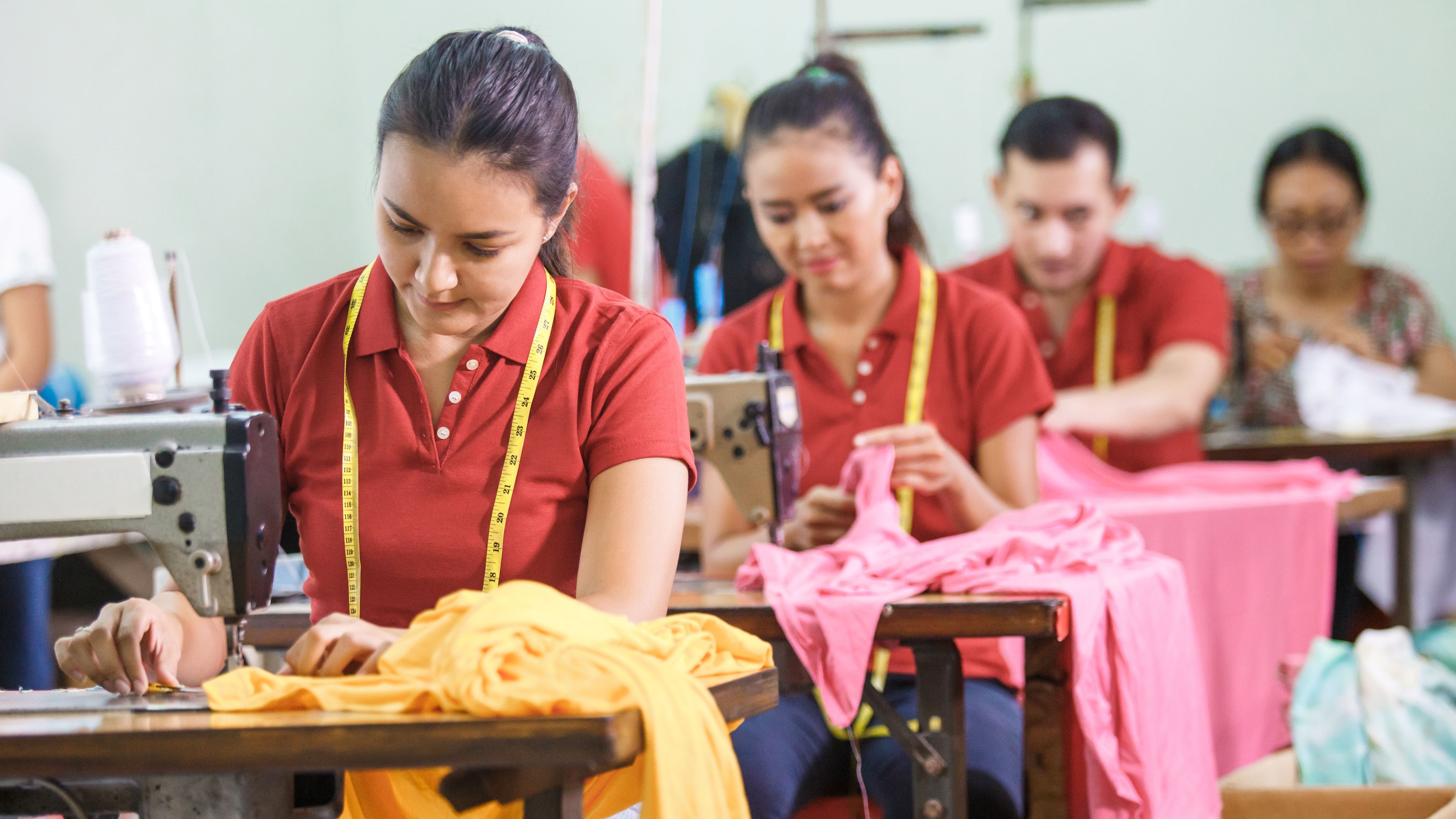 Workers in a sewing workshop are focused on stitching and handling colorful fabric. They are wearing red shirts and yellow measuring tapes around their necks. Sewing machines and fabric are visible on the shared workspace.