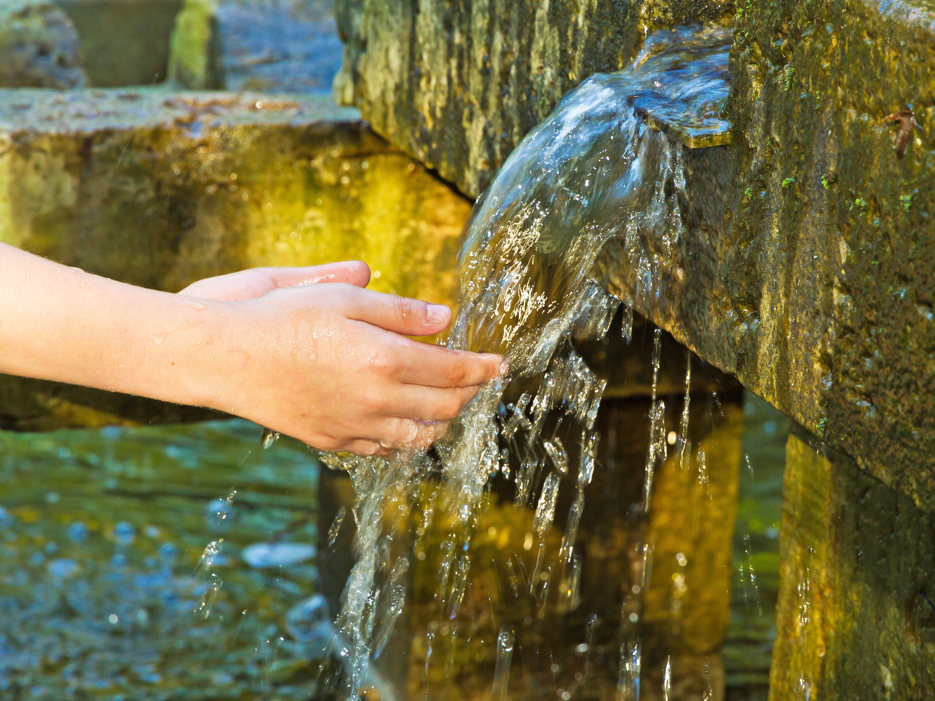 A close-up of a hand reaching out to catch flowing water from a stone fountain. The water sparkles in the sunlight, and moss and greenery cover parts of the fountain. A tranquil scene with natural and rustic elements.