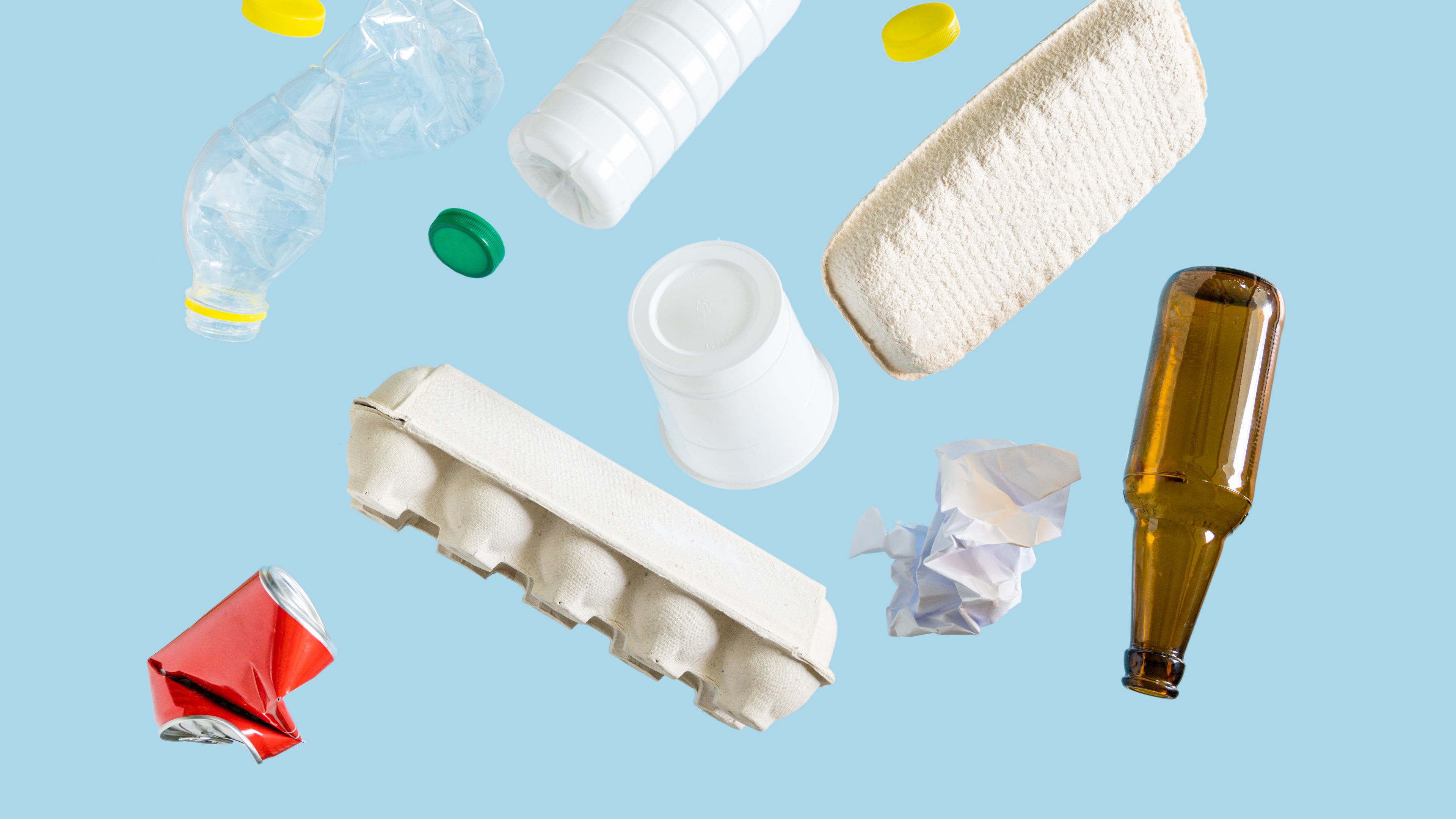 Various recyclable materials, including a plastic bottle, aluminum can, glass bottle, paper egg carton, crumpled paper, and bottle caps, are scattered against a light blue background. The arrangement showcases different types of materials often collected for recycling.