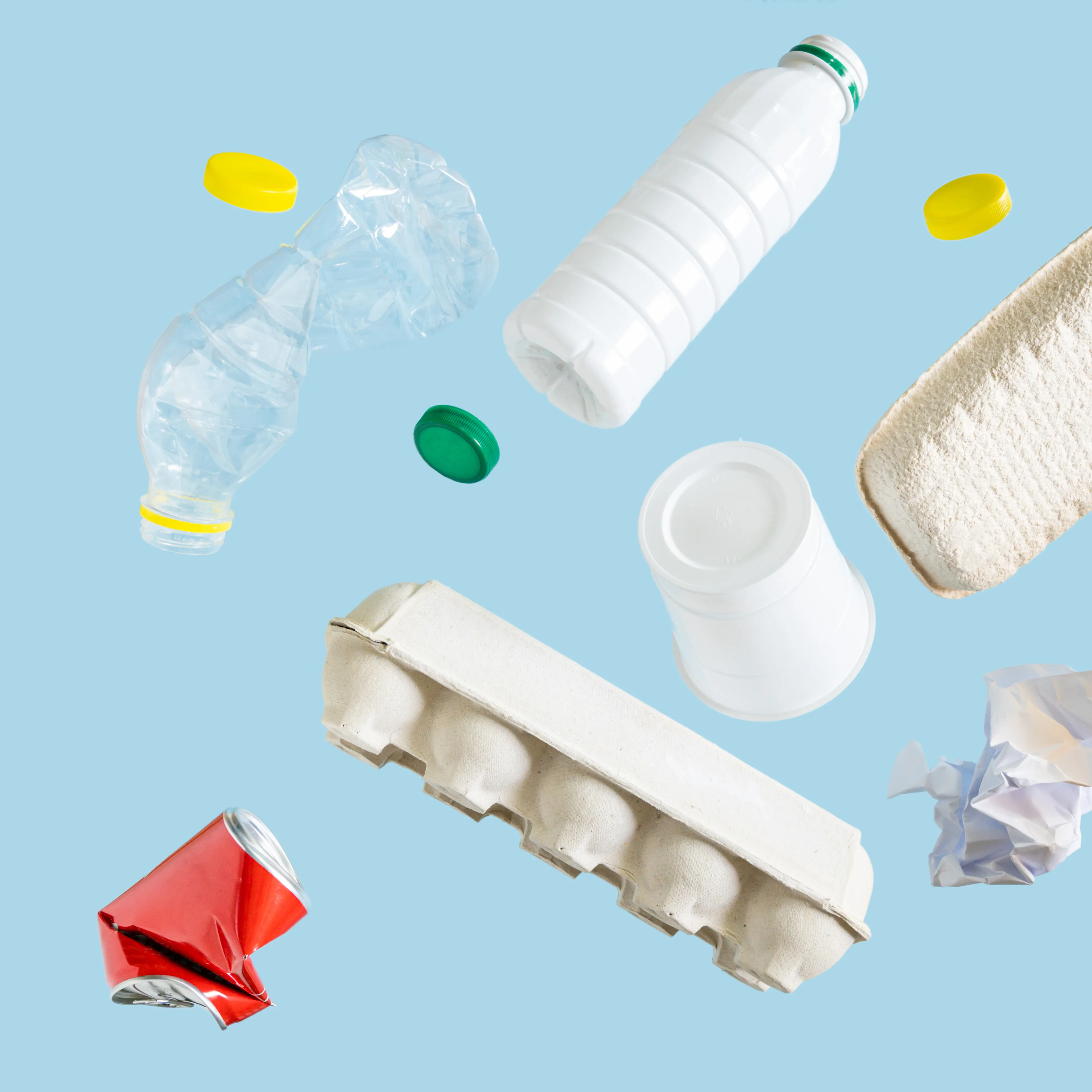 Various recyclable materials, including a plastic bottle, aluminum can, glass bottle, paper egg carton, crumpled paper, and bottle caps, are scattered against a light blue background. The arrangement showcases different types of materials often collected for recycling.