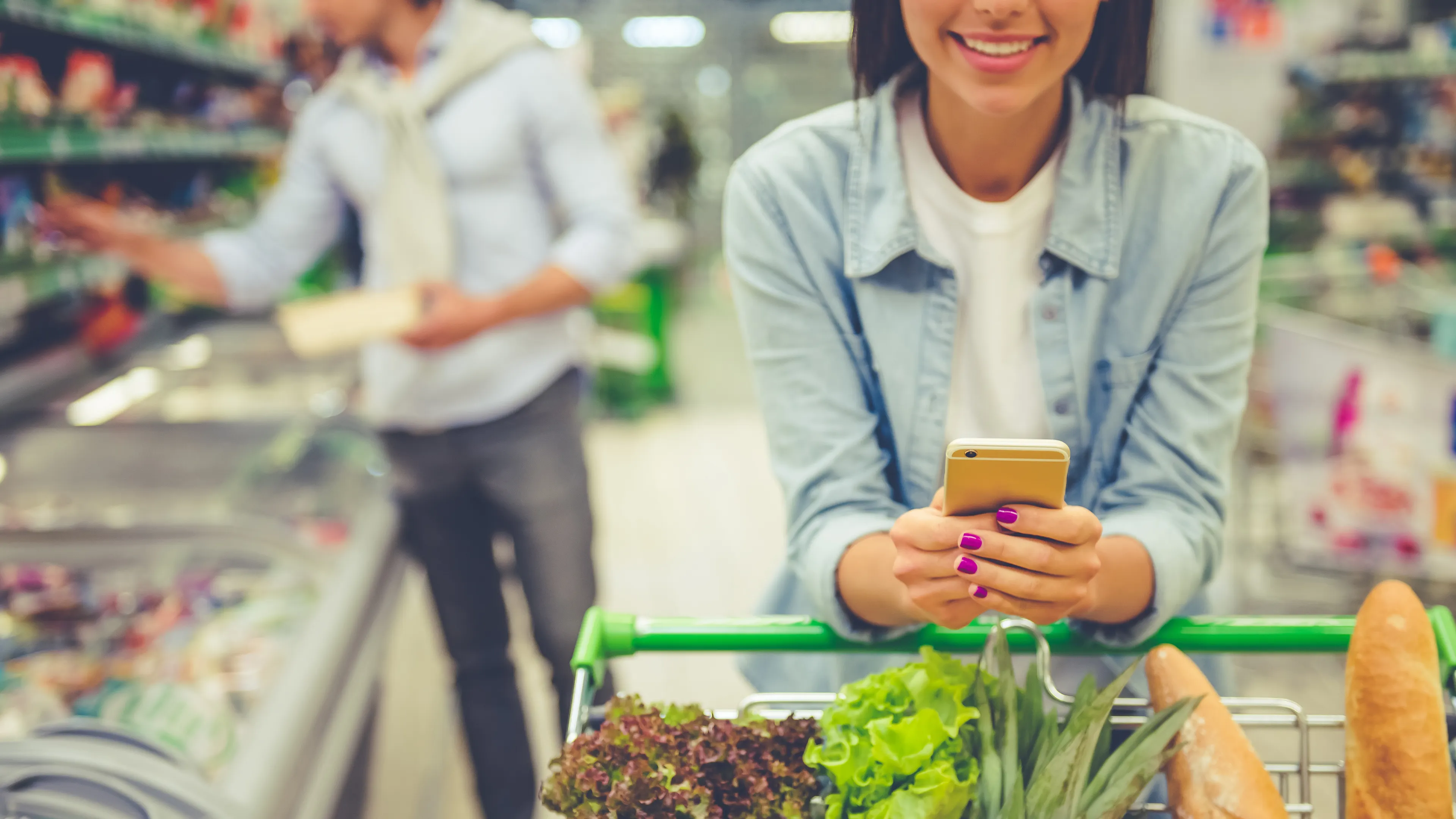 A woman is smiling while holding her phone and leaning on a shopping cart filled with fresh produce, including lettuce, pineapple, carrots, and baguettes. A man in the background is browsing items on a shelf in a grocery store. The setting appears bright and organized.