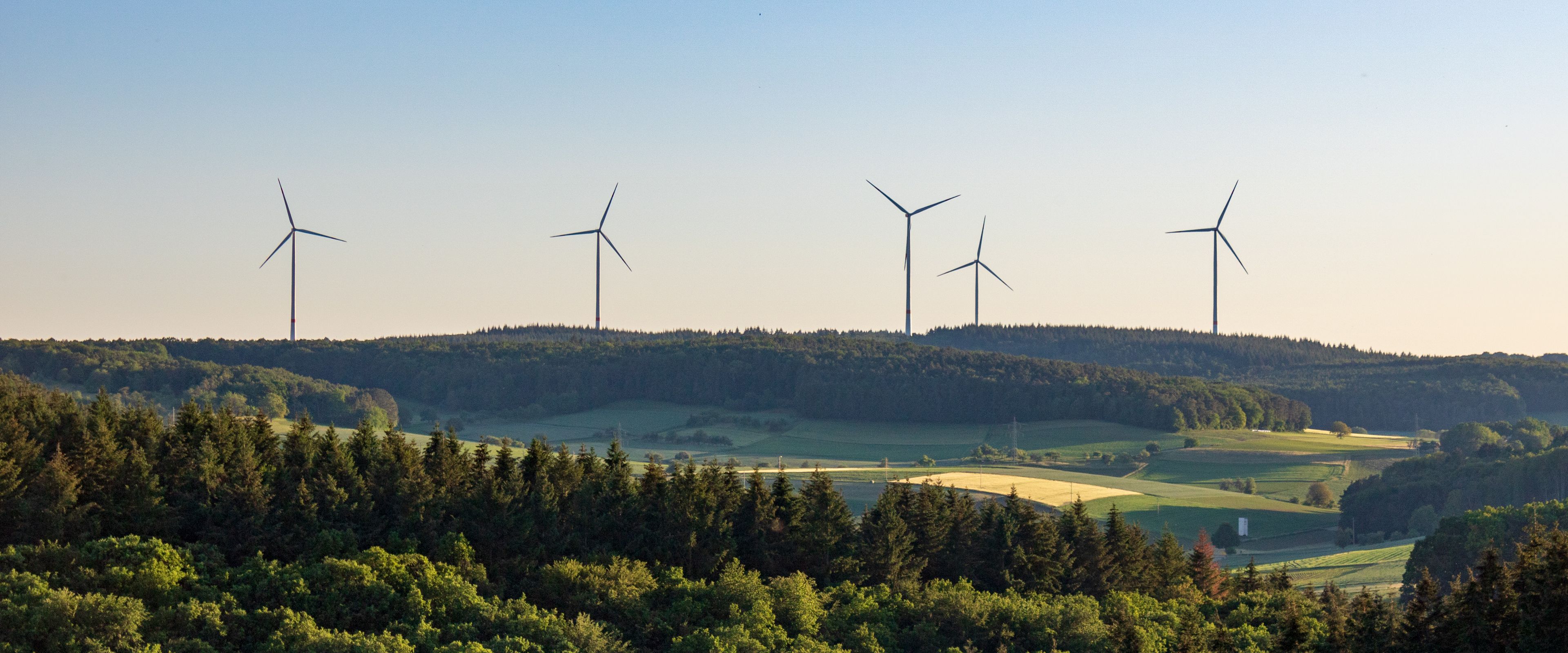 A landscape featuring several wind turbines on a hill surrounded by lush greenery and forests. The sky is clear and blue, creating a serene atmosphere. Rolling fields are visible between the forested areas.