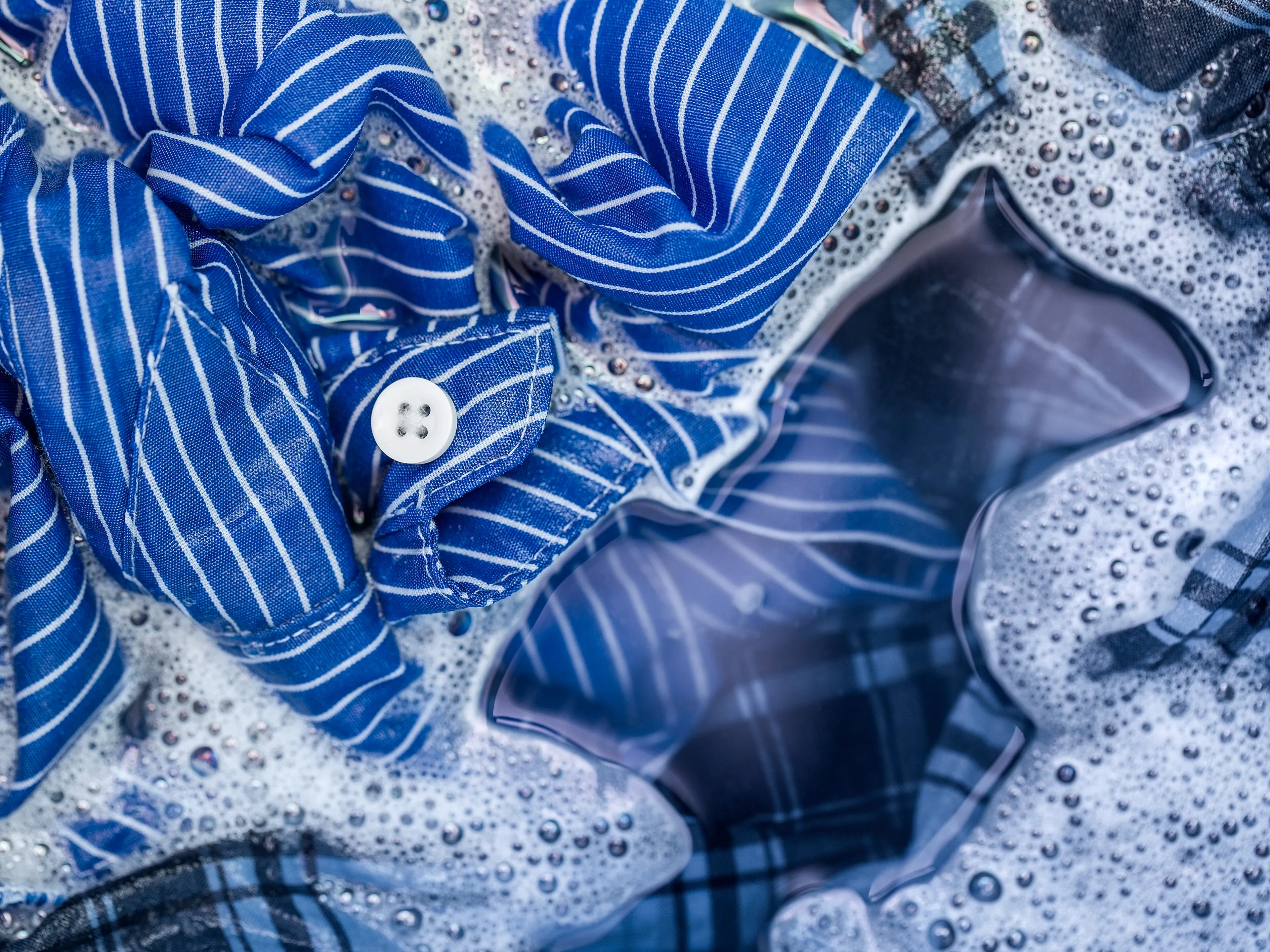 Blue striped and plaid fabrics, including a blue shirt with a white button, soaking in soapy water. White suds and reflections of light are visible on the surface. The image suggests clothes being washed.