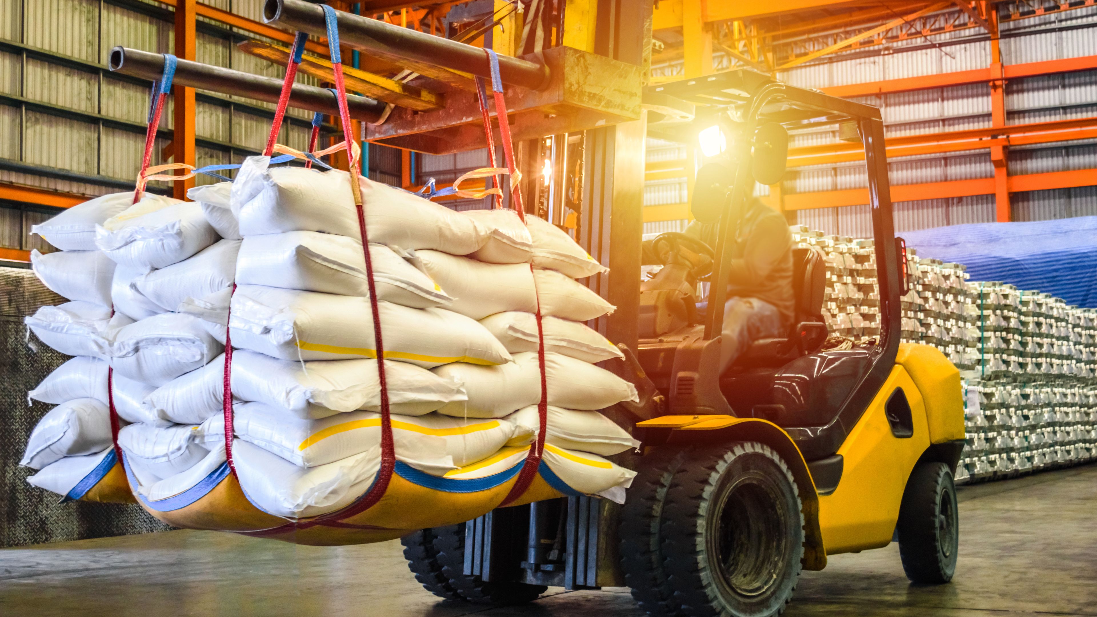 A forklift transports a large stack of white bags secured with straps inside a warehouse. The area is brightly lit, with shelves in the background holding more bags. The scene depicts an industrial storage or logistics operation.