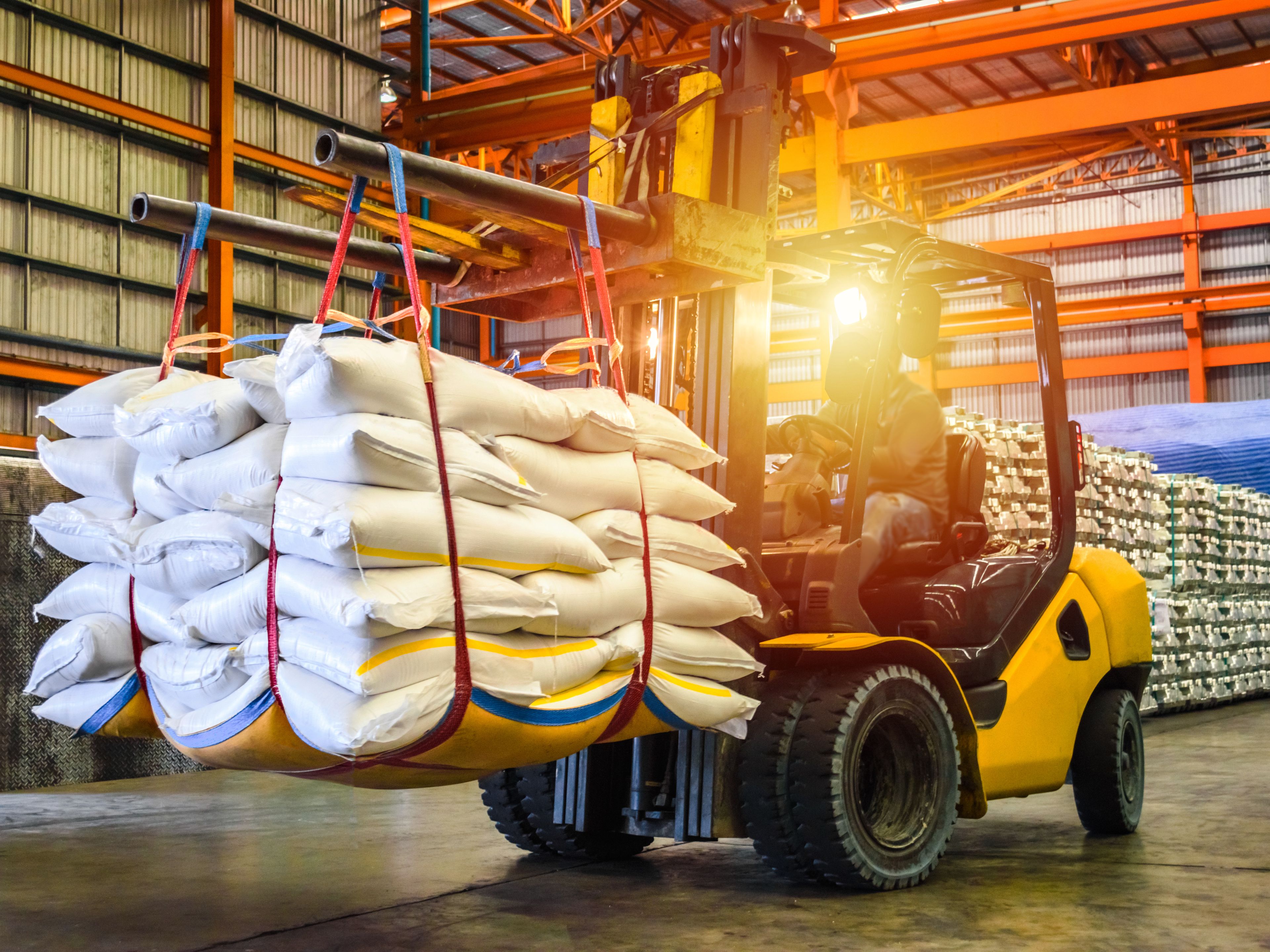A forklift transports a large stack of white bags secured with straps inside a warehouse. The area is brightly lit, with shelves in the background holding more bags. The scene depicts an industrial storage or logistics operation.