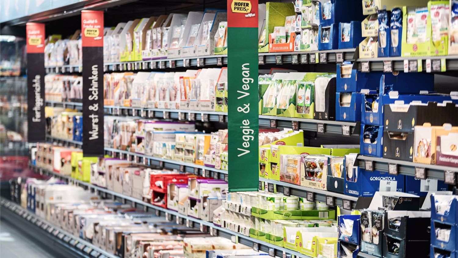 A HOFER supermarket aisle filled with various packaged products, organized neatly on shelves. A green sign with "Veggie & Vegan" hangs prominently, indicating a section dedicated to vegetarian and vegan food options. Bright lighting illuminates the well-stocked shelves.