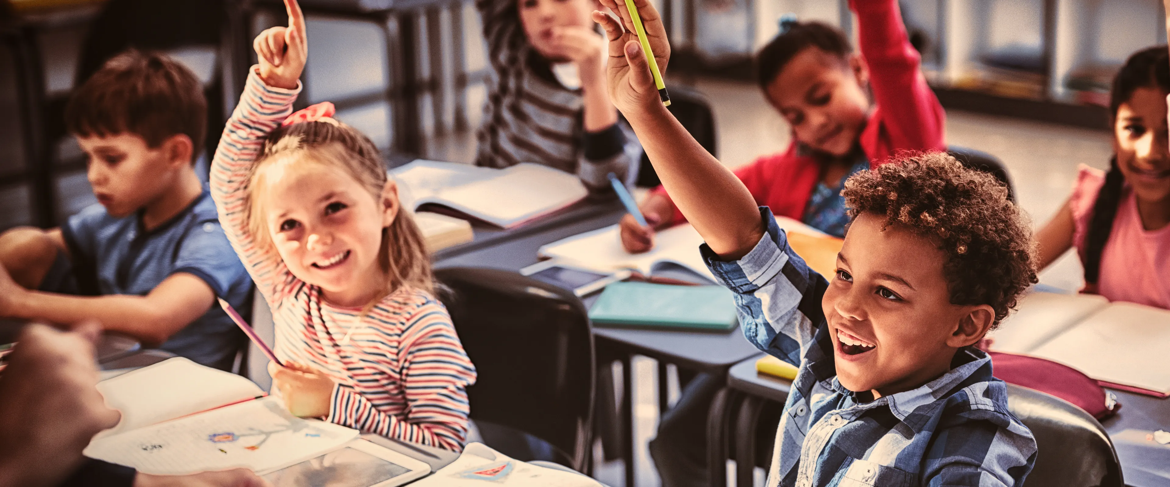 A classroom of young children seated at desks, enthusiastically raising their hands to answer a question. The students are diverse and smiling, with school supplies and books visible on their desks. The atmosphere appears lively and engaging.