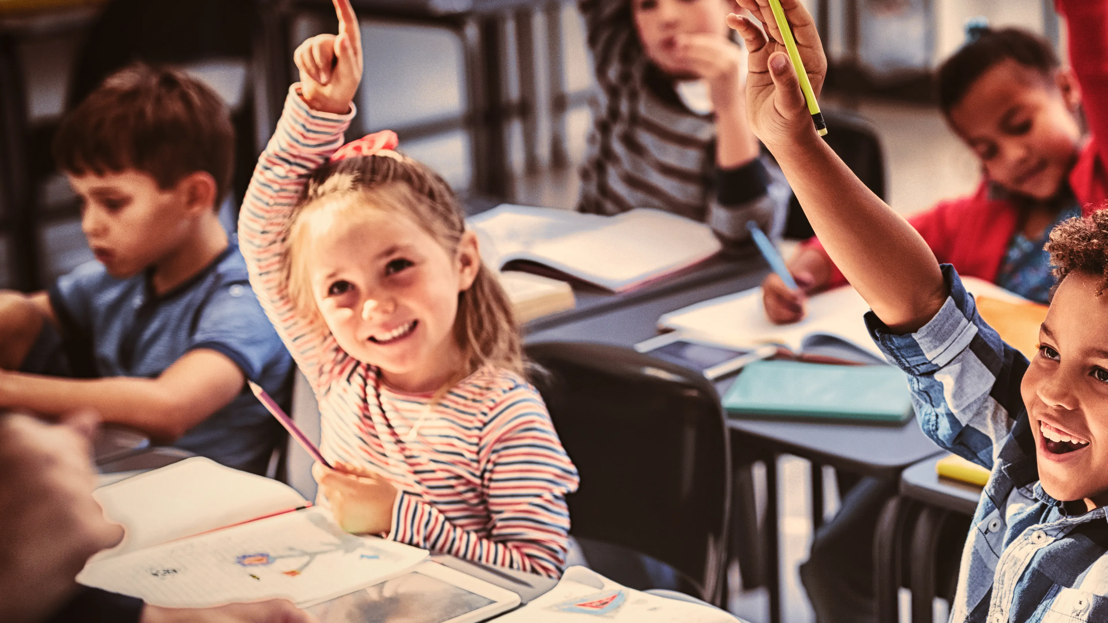 A classroom of young children seated at desks, enthusiastically raising their hands to answer a question. The students are diverse and smiling, with school supplies and books visible on their desks. The atmosphere appears lively and engaging.