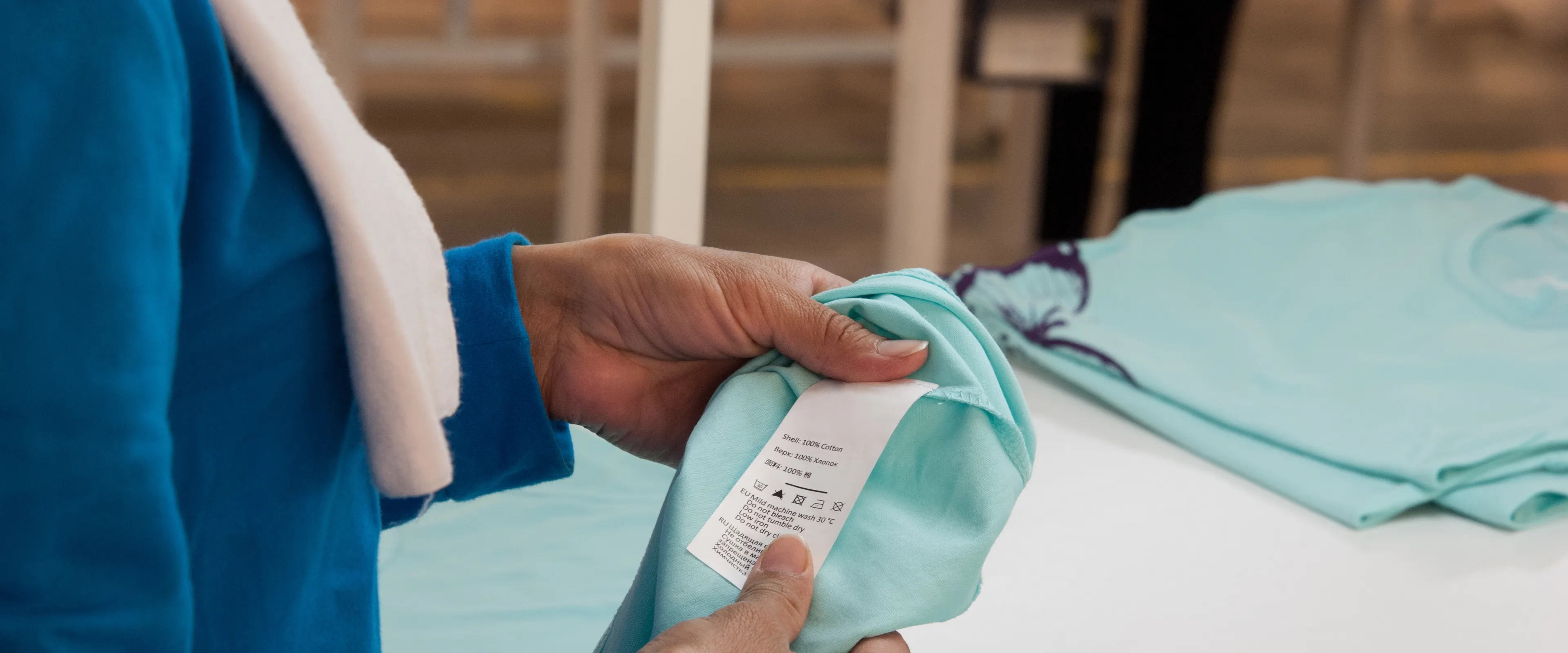 A person in a blue long-sleeve shirt is examining the label of a light turquoise t-shirt. The label displays care instructions and fabric details. Other similar t-shirts are laid on a white table in the background.