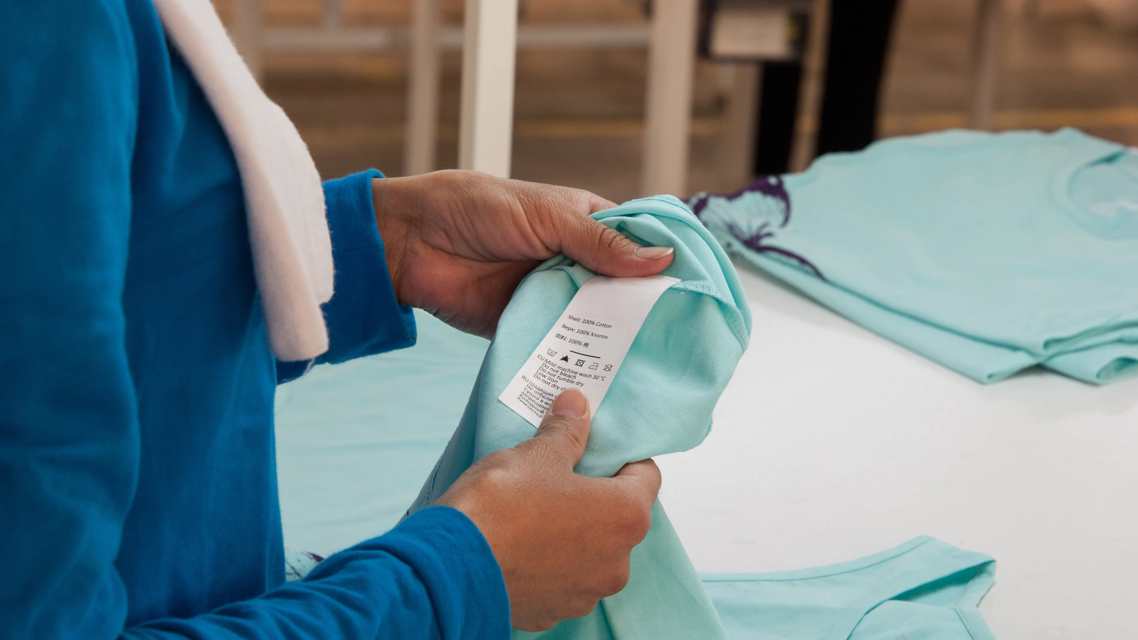 A person in a blue long-sleeve shirt is examining the label of a light turquoise t-shirt. The label displays care instructions and fabric details. Other similar t-shirts are laid on a white table in the background.