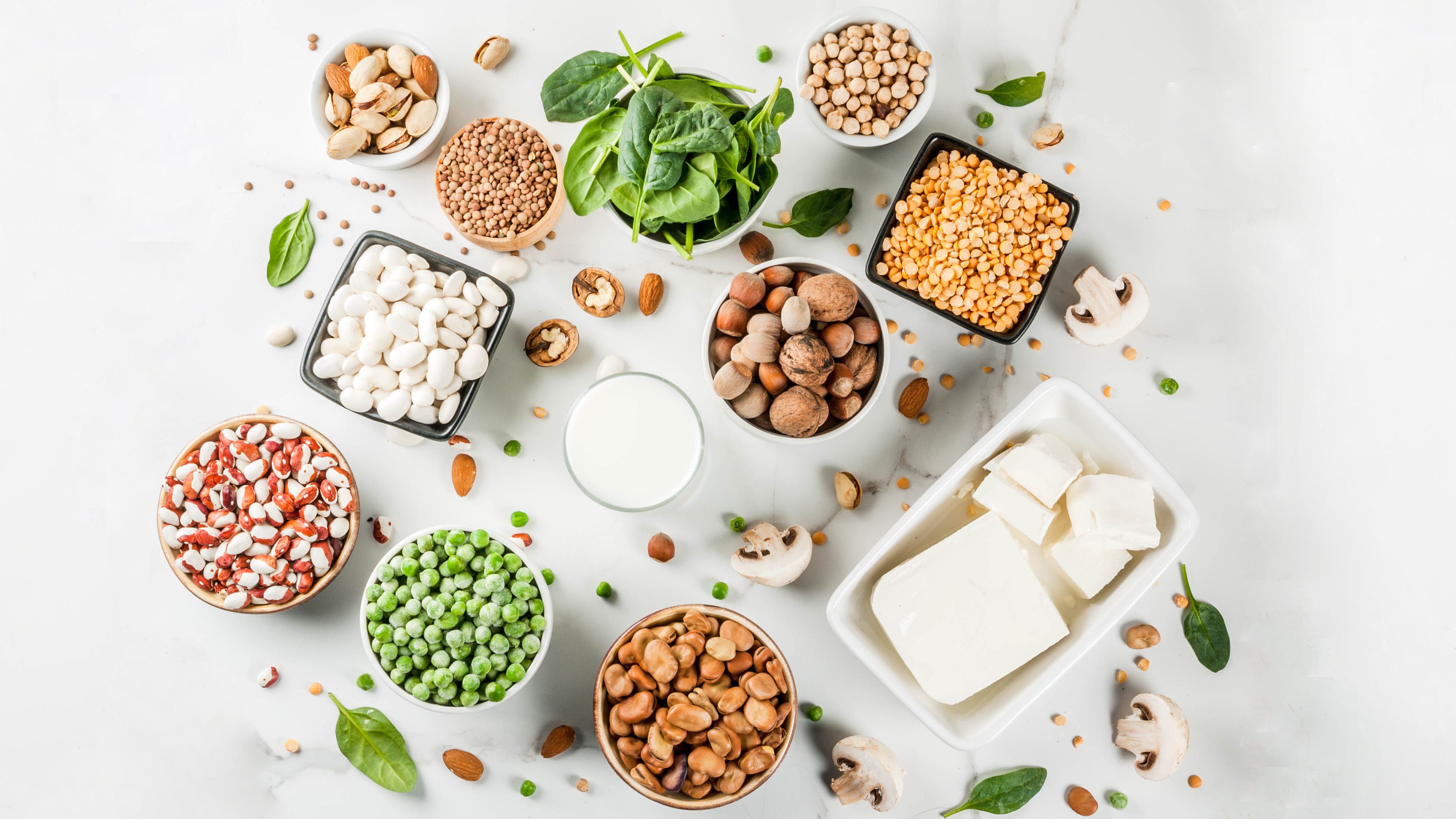An assortment of plant-based protein sources displayed on a white surface, including tofu, legumes, nuts, peas, lentils, beans, spinach, and mushrooms. A small bowl of plant-based milk is also placed amongst the ingredients. Scattered leaves and nuts add a natural touch to the arrangement.