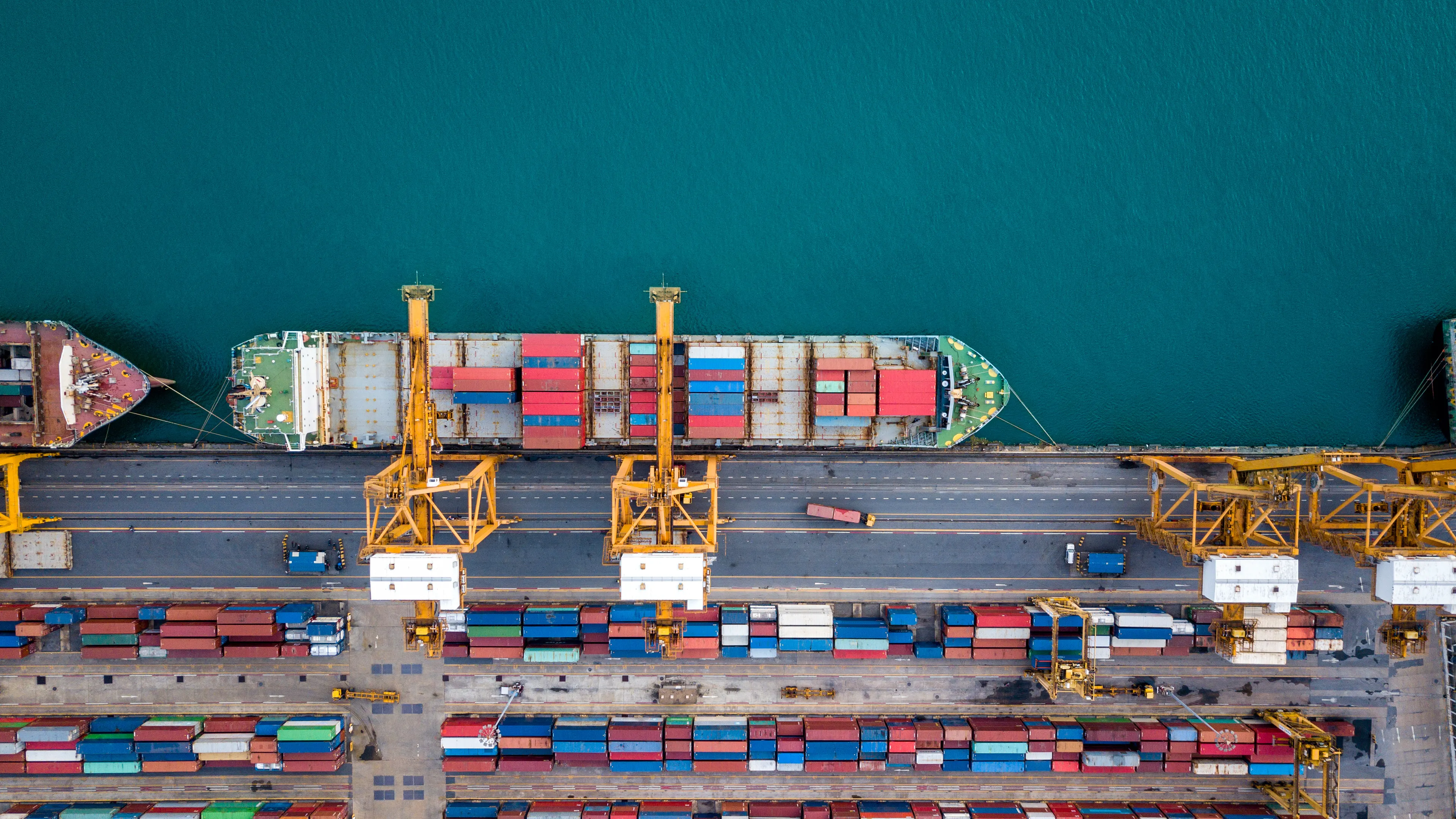 An aerial view of a busy shipping port, showing large cargo ships docked alongside. Rows of colorful shipping containers are stacked neatly, with cranes positioned for loading and unloading. The turquoise water of the harbor contrasts with the vibrant colors of the containers.