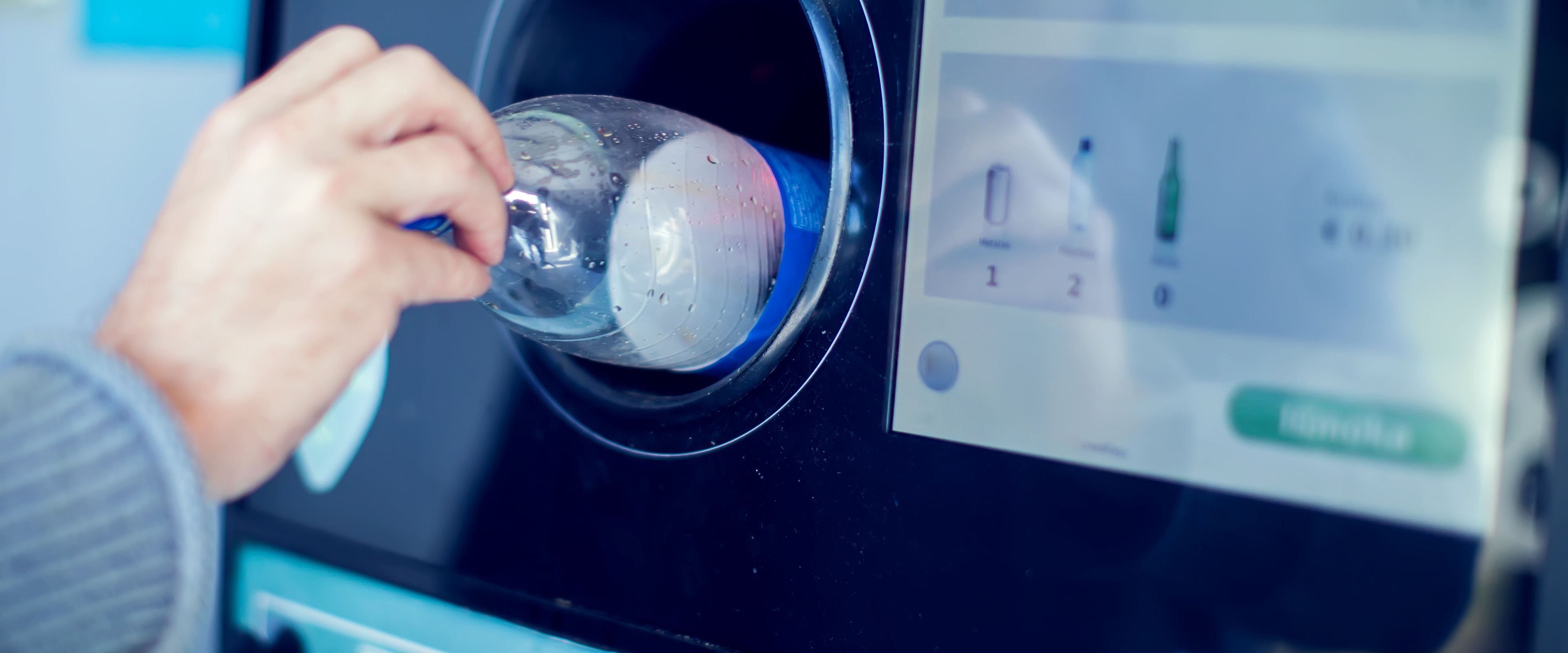 A person inserting a plastic bottle into a reverse vending machine designed for recycling. The machine has a display screen showing recycling instructions and options.