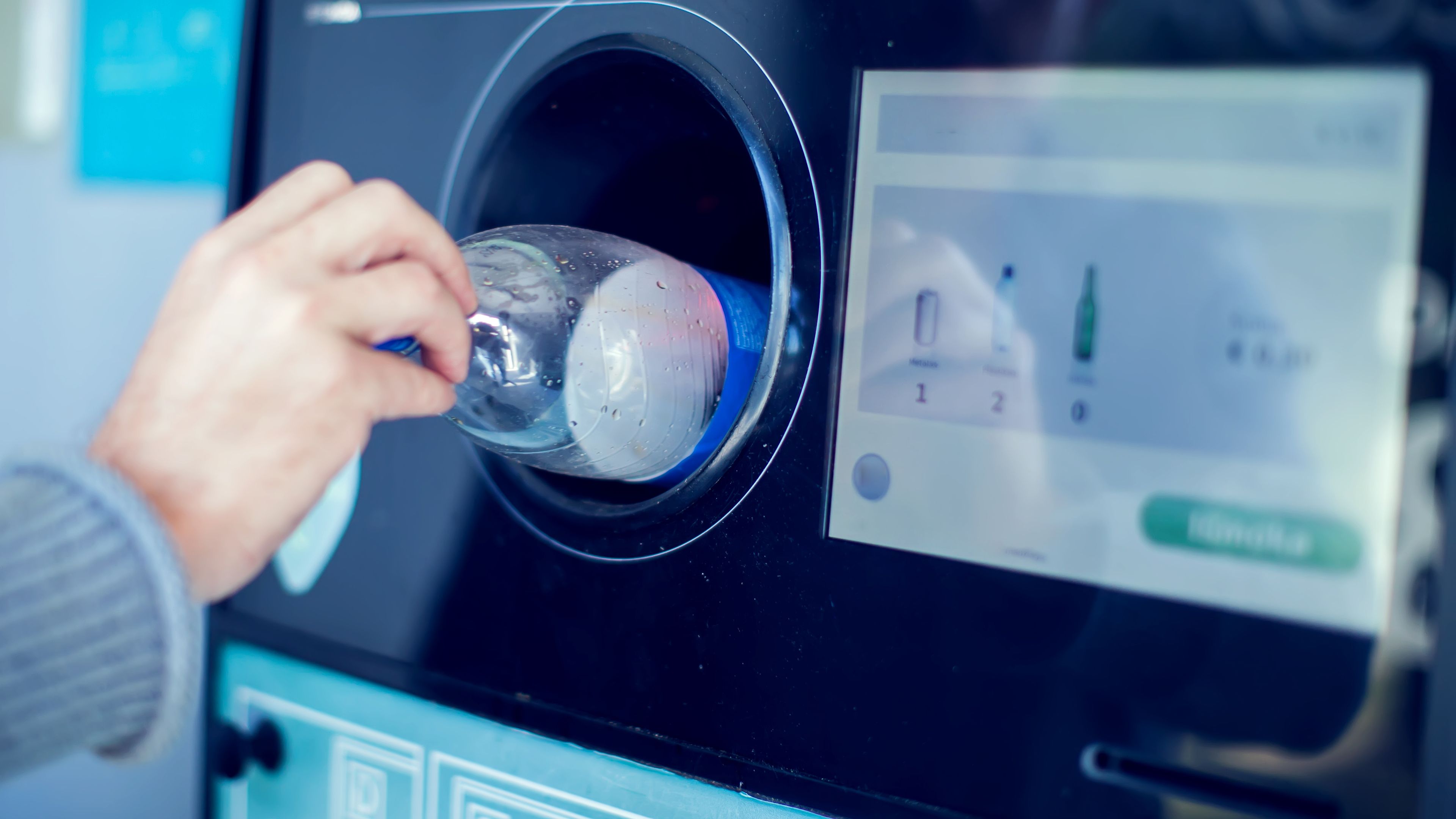A person inserting a plastic bottle into a reverse vending machine designed for recycling. The machine has a display screen showing recycling instructions and options.