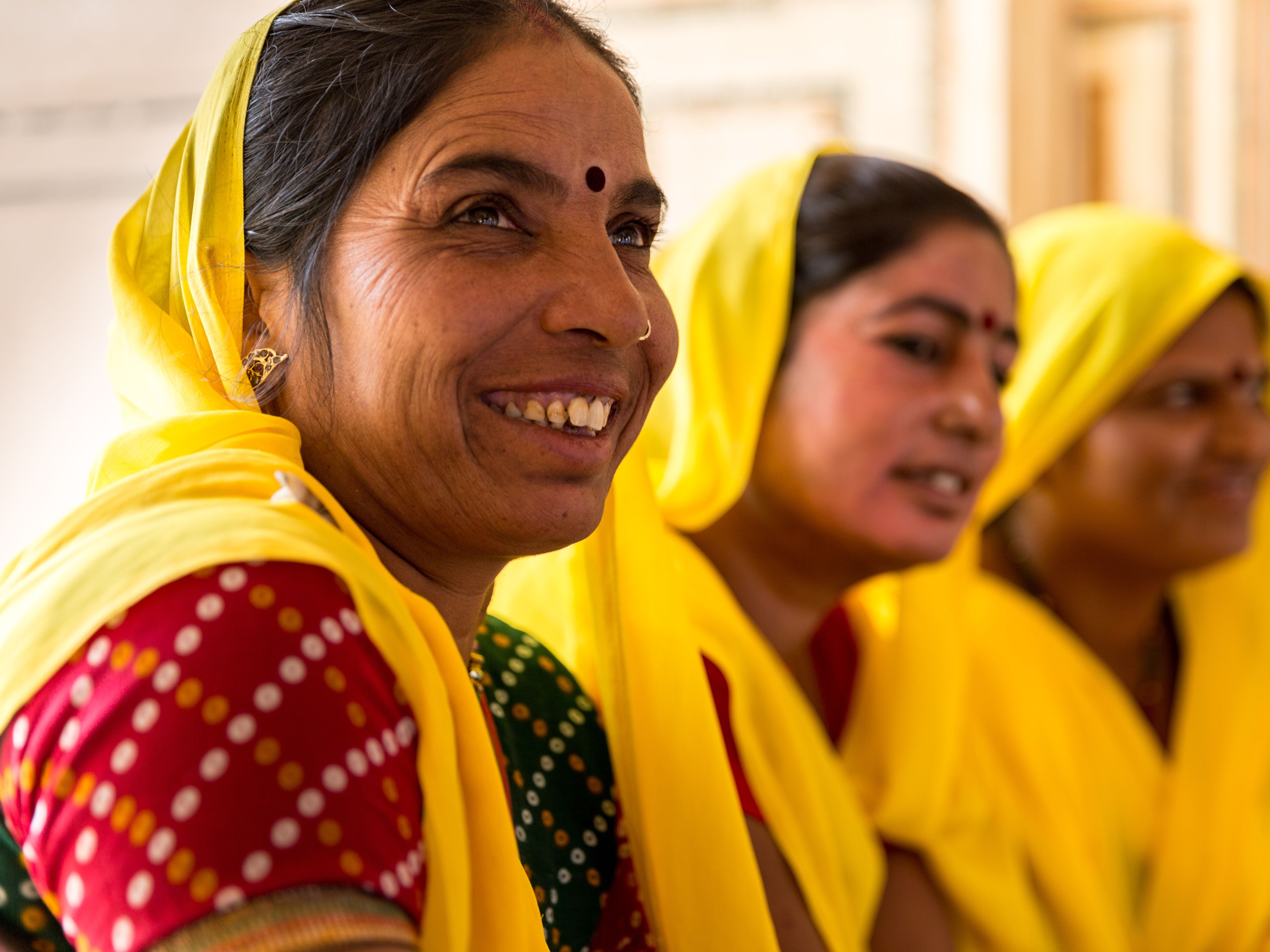 Three women dressed in vibrant yellow sarees and traditional attire are gathered together, smiling and conversing. The woman in the foreground has a joyful expression and a red bindi on her forehead. The background is softly blurred, highlighting their interaction.