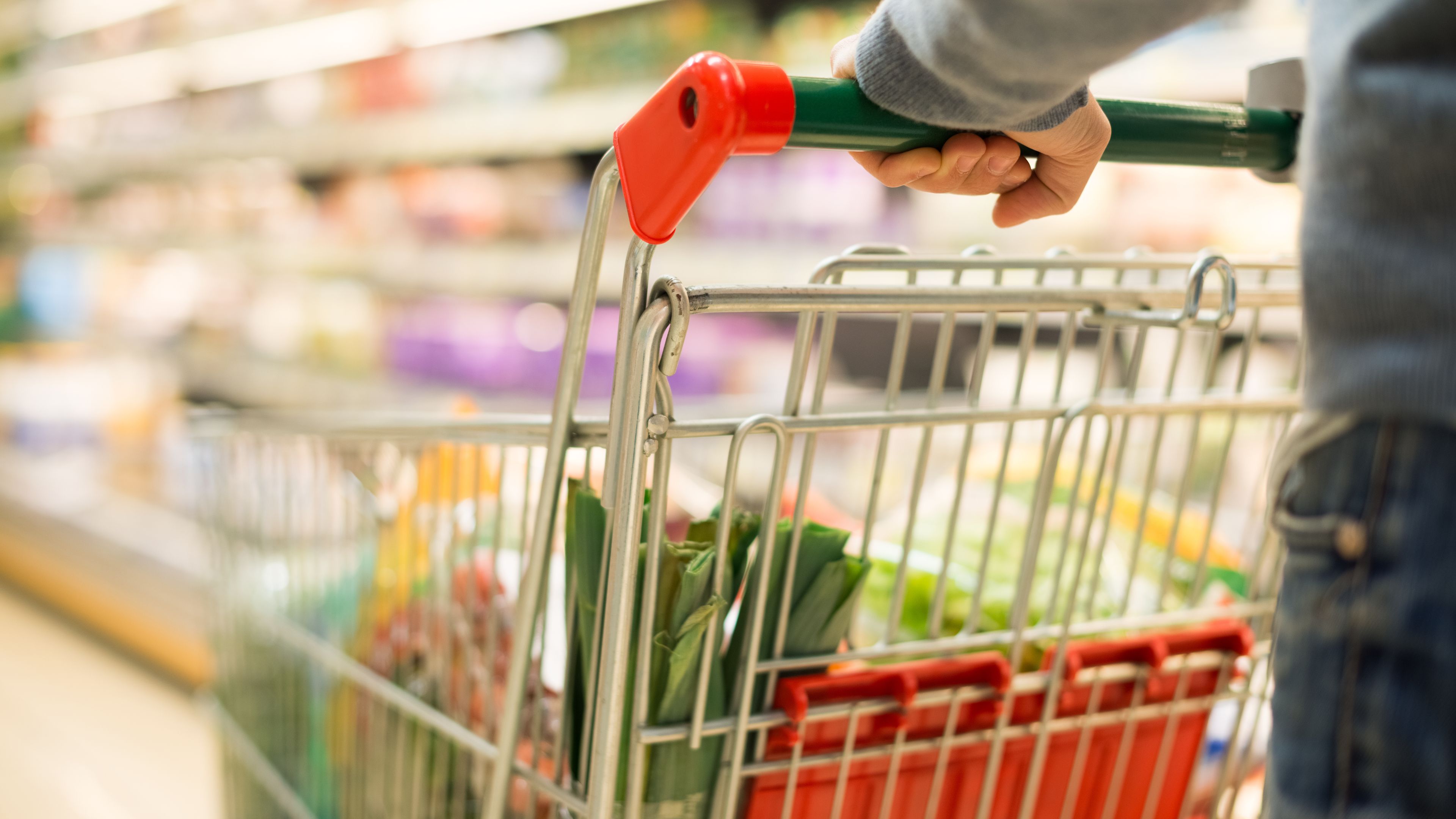 A person pushes a shopping cart filled with groceries in a supermarket aisle. The cart contains packaged goods and fresh produce. Shelves stocked with various products are visible in the background.