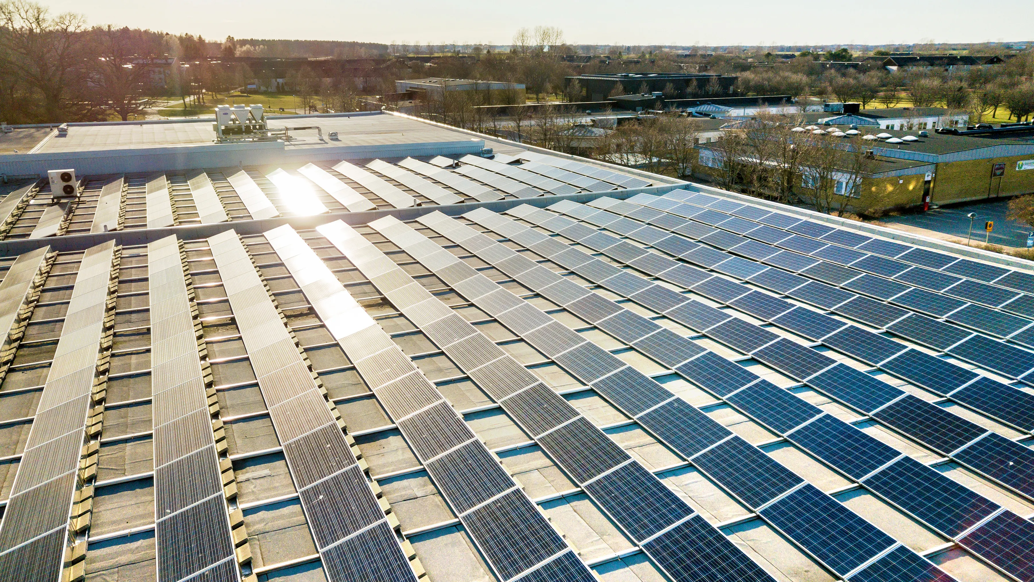 A large rooftop covered entirely with solar panels, glinting under the sunlight. The background shows open spaces and distant buildings. Some trees are visible beyond the structures.
