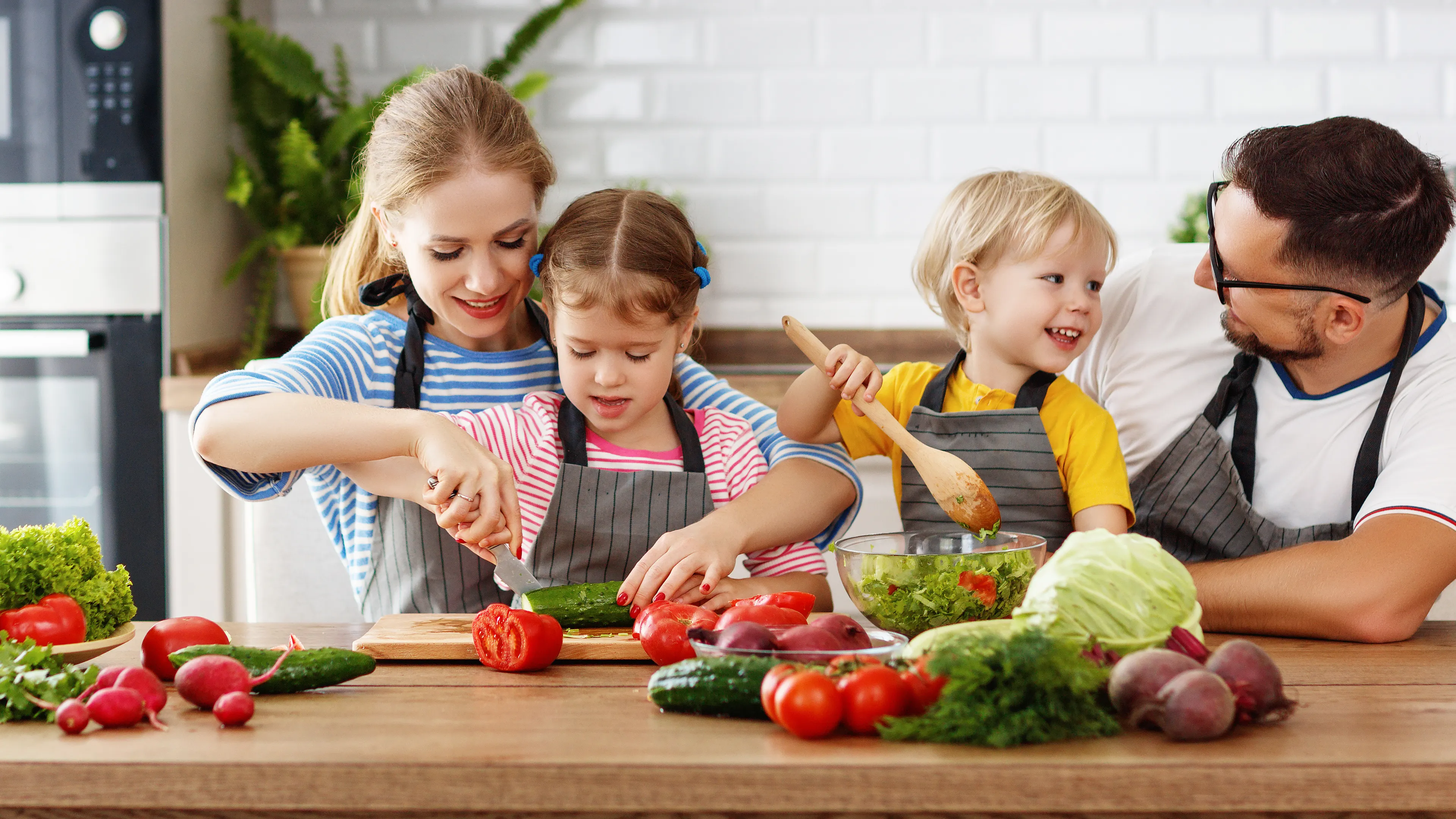 A family of four, two adults and two children, is gathered in a bright kitchen preparing food. They are smiling and wearing aprons while chopping vegetables and mixing ingredients. Fresh produce, such as tomatoes, cucumbers, and lettuce, is spread across the wooden countertop.