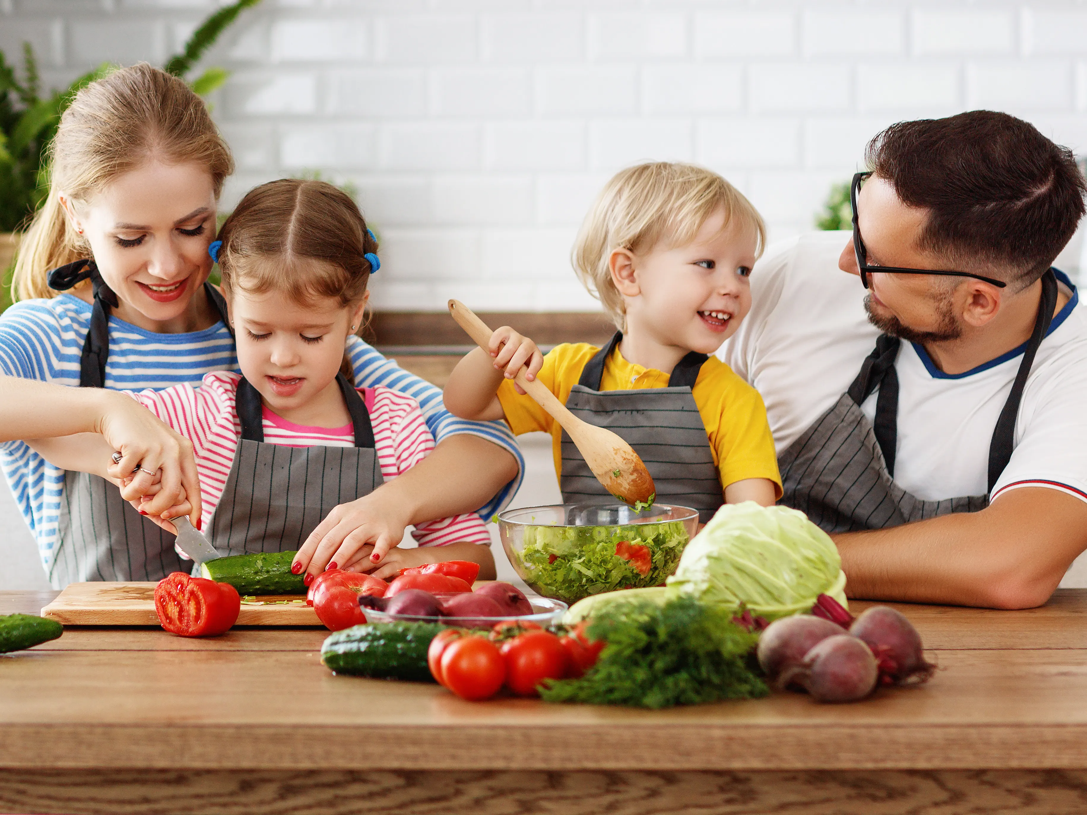 A family of four, two adults and two children, is gathered in a bright kitchen preparing food. They are smiling and wearing aprons while chopping vegetables and mixing ingredients. Fresh produce, such as tomatoes, cucumbers, and lettuce, is spread across the wooden countertop.