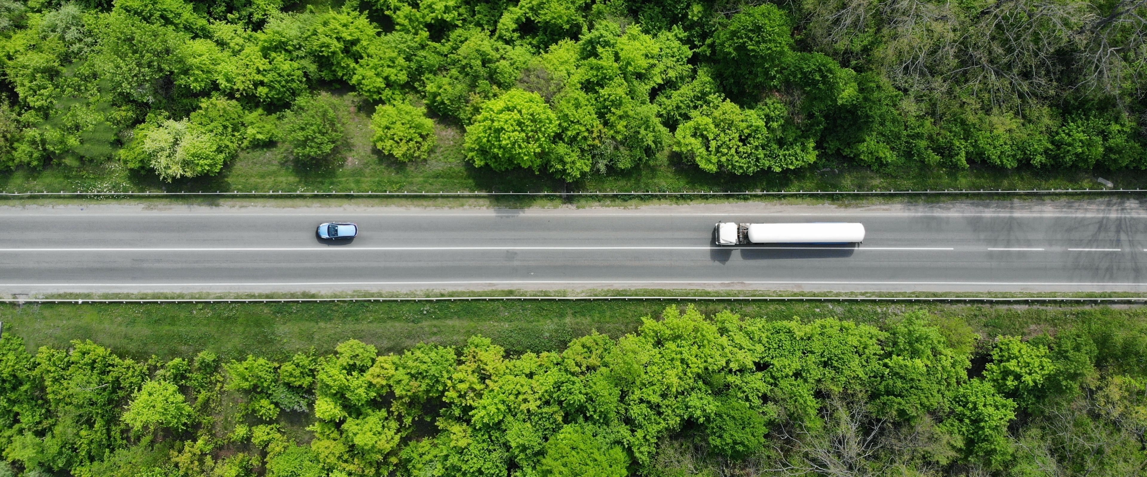 Aerial view of a two-lane road surrounded by lush green trees and vegetation. A blue car and a white semi-truck are traveling in opposite directions. The scene showcases a peaceful countryside setting.