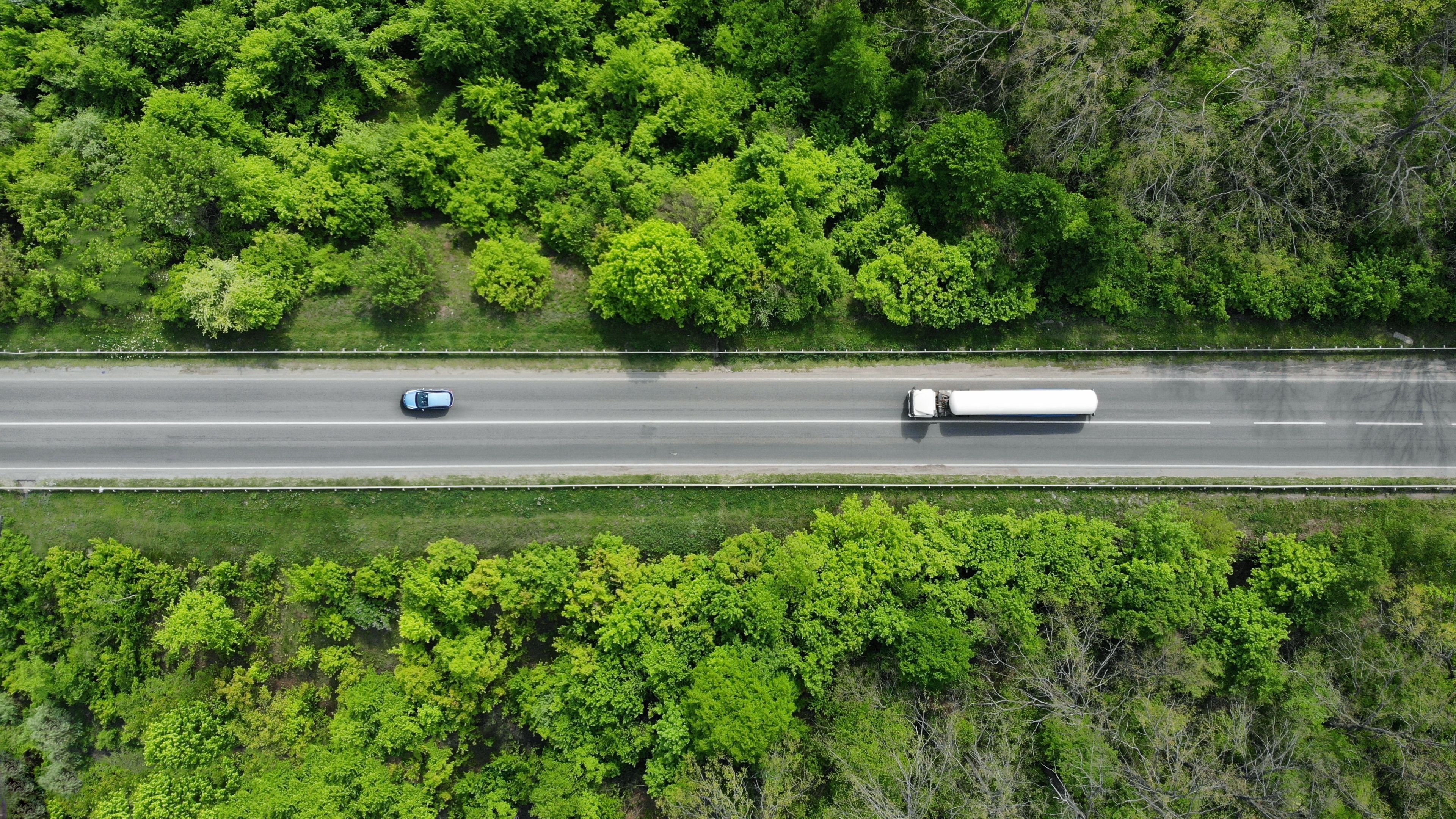 Aerial view of a two-lane road surrounded by lush green trees and vegetation. A blue car and a white semi-truck are traveling in opposite directions. The scene showcases a peaceful countryside setting.