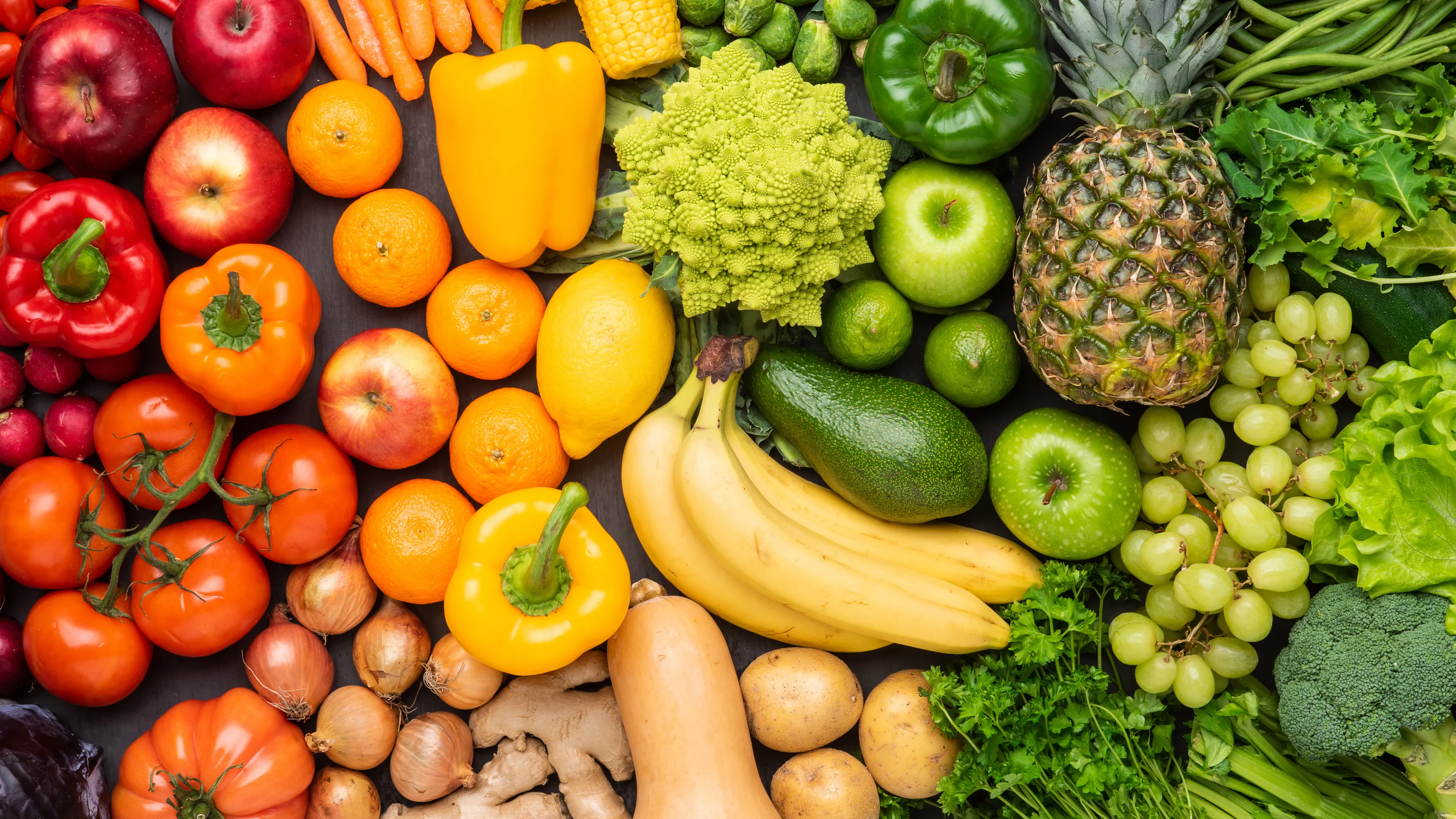 A colorful assortment of fresh fruits and vegetables is displayed, including apples, bell peppers, bananas, tomatoes, carrots, corn, and a pineapple. The produce is arranged neatly, showcasing a variety of colors and textures. Green leafy vegetables and other fresh items are also part of the vibrant arrangement.