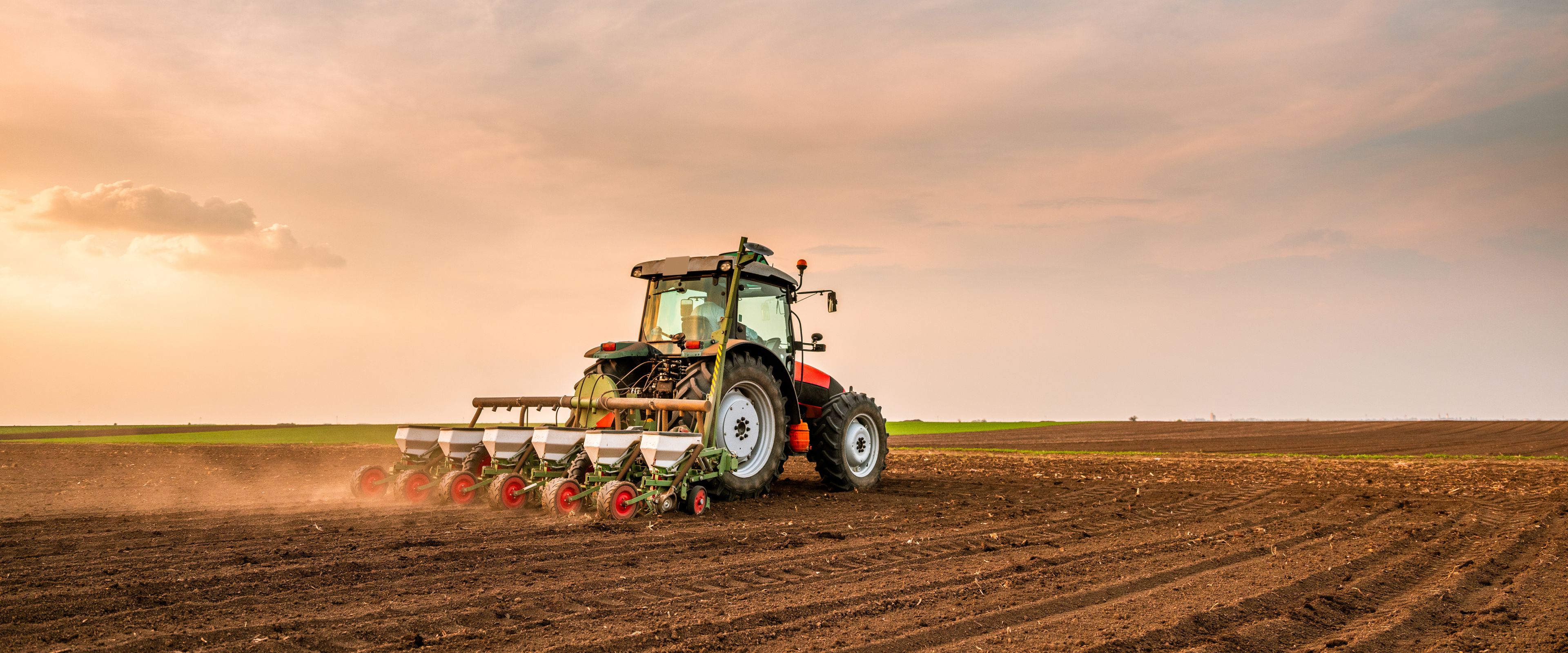 A tractor is plowing a large, empty field at sunset under an expansive sky. The soil appears freshly tilled, and the sun casts a soft, warm light across the landscape.