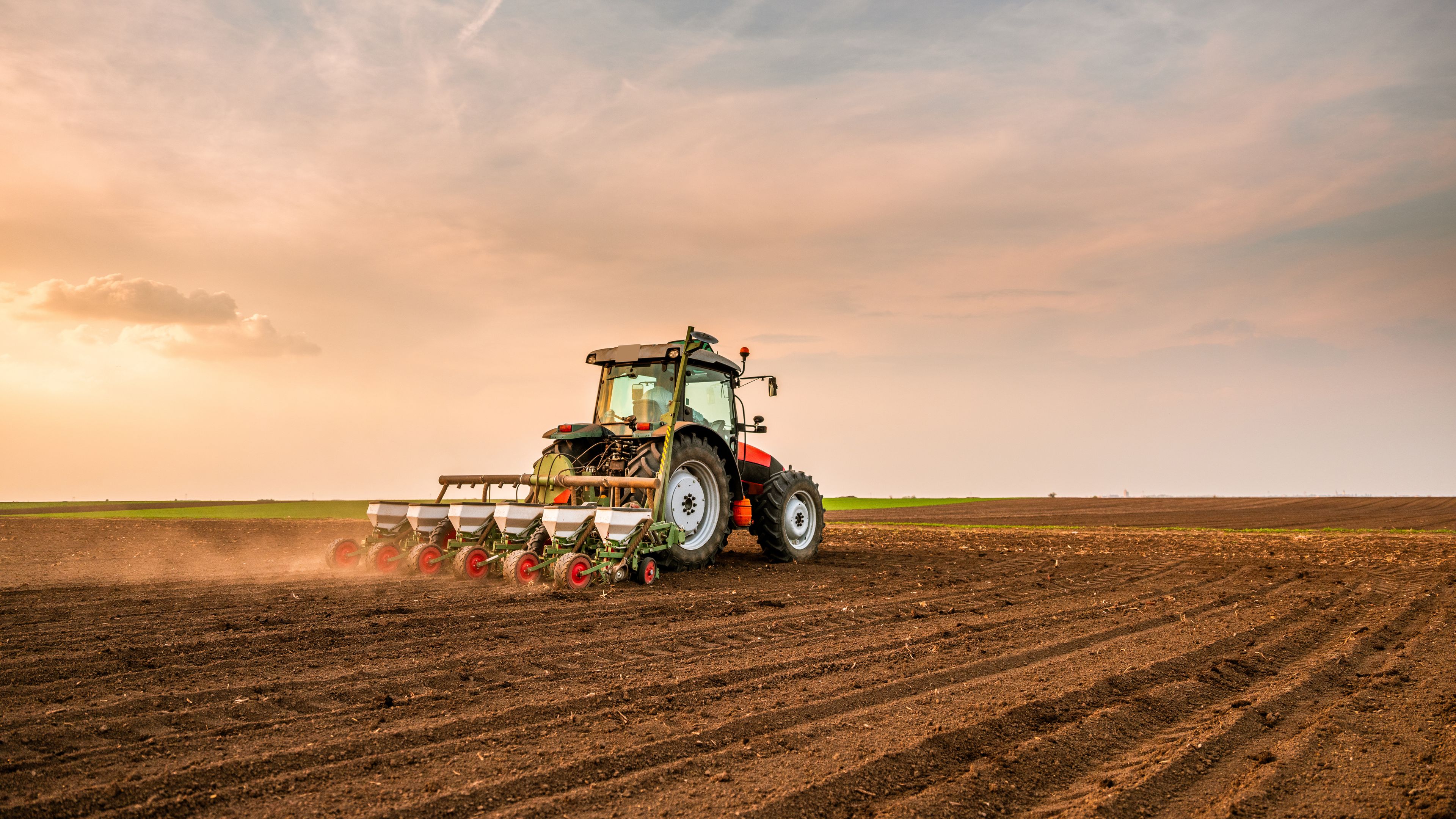 A tractor is plowing a large, empty field at sunset under an expansive sky. The soil appears freshly tilled, and the sun casts a soft, warm light across the landscape.